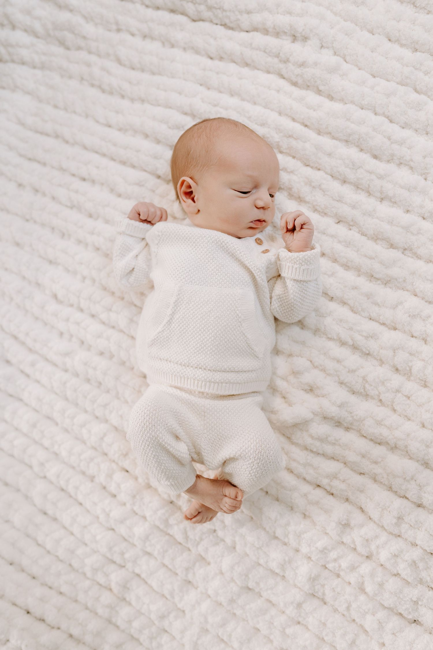 Peaceful newborn baby sleeping on a white textured blanket wearing a cream knit outfit.
