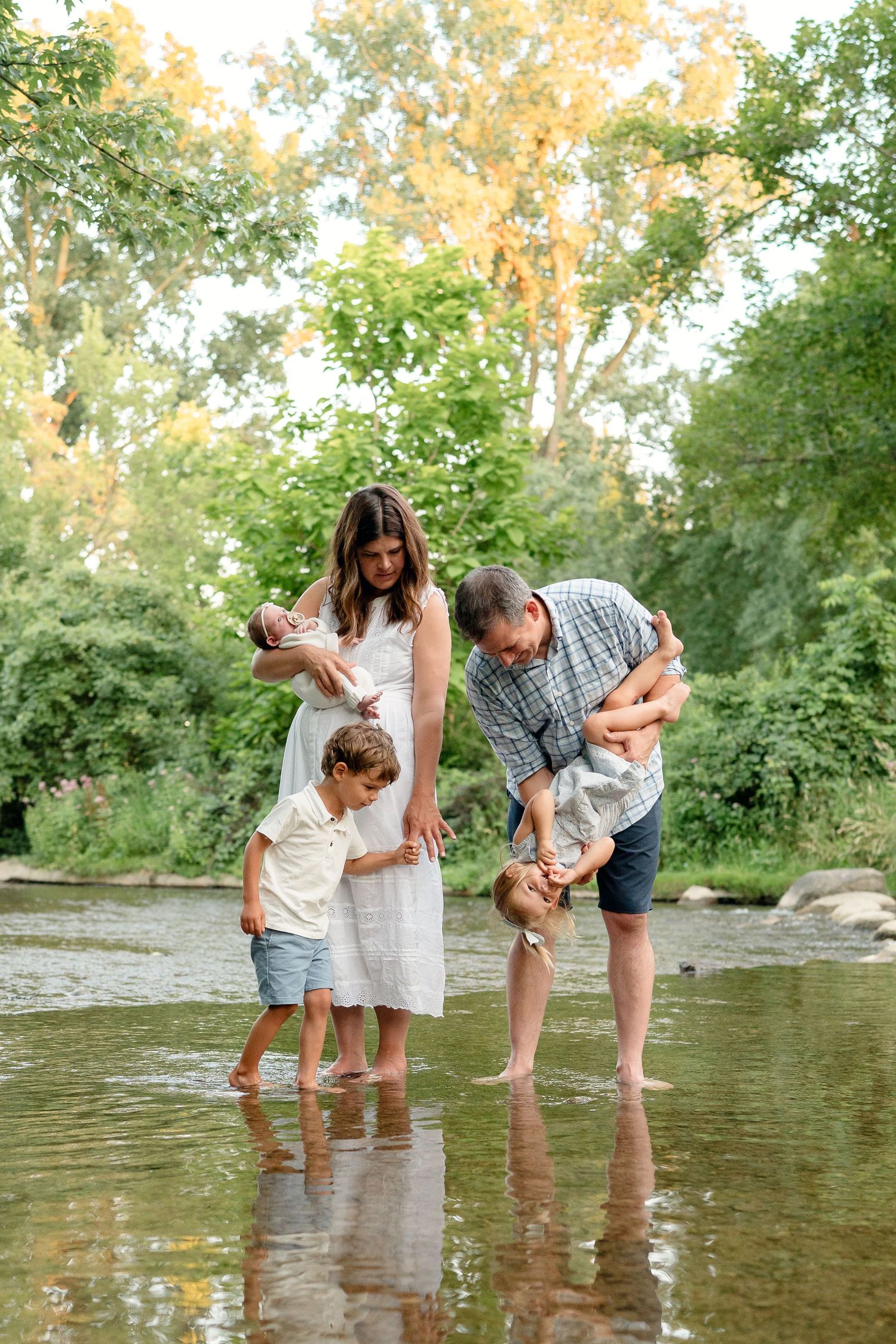 Family of four wading in a shallow creek surrounded by lush green trees, parents holding children outdoors.