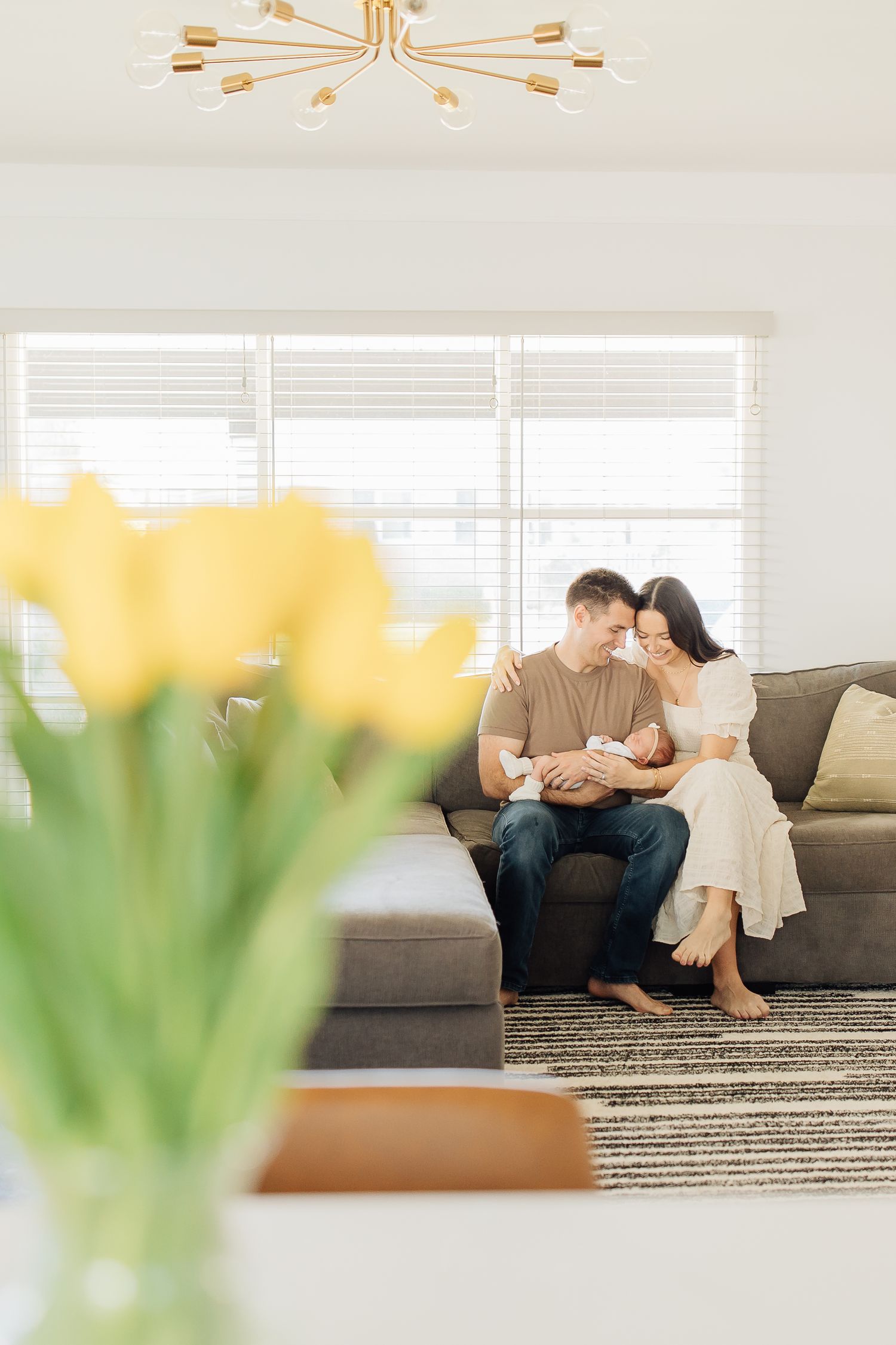 A couple sits on a couch gazing at their newborn with yellow tulips in the foreground.