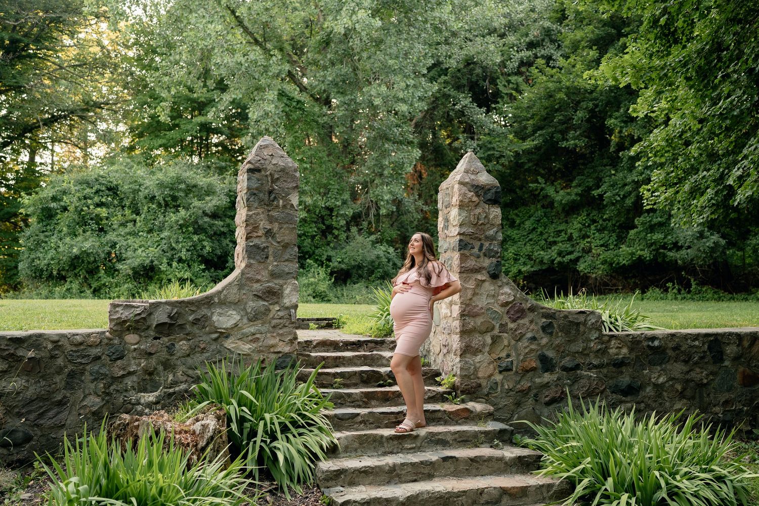 Pregnant woman in a pink dress posing gracefully on stone steps between rustic pillars in a lush garden setting.