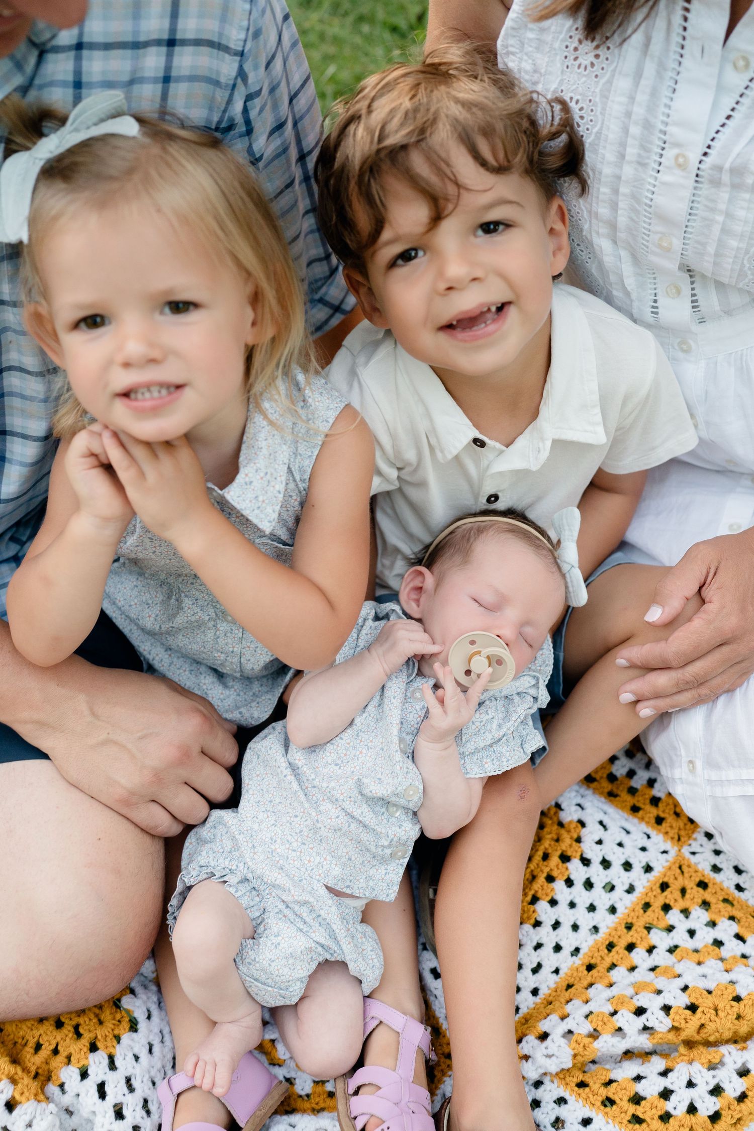 Two young siblings smile outdoors holding a sleeping newborn baby on a yellow blanket during a family photo session.