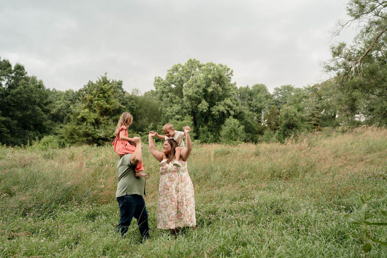 Family playing together in a lush green meadow with trees in background on a cloudy day.