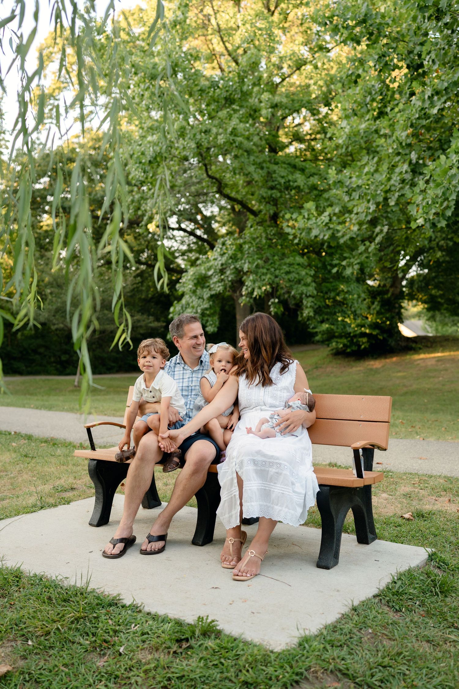 Family of four with newborn sitting on a park bench surrounded by lush green trees on a sunny day.