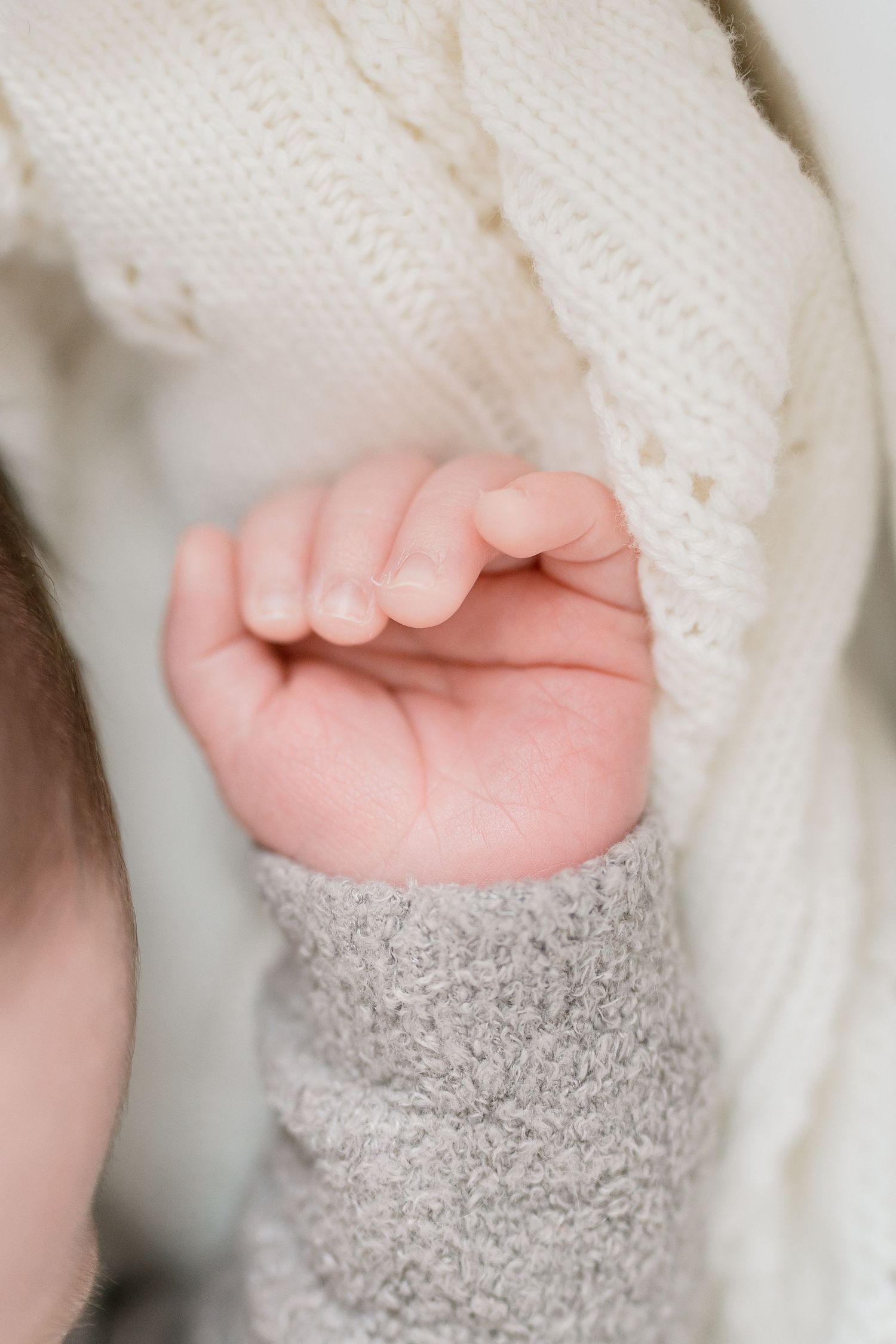 Close-up of a newborn baby's tiny hand resting against a soft white knit blanket and gray sweater sleeve.