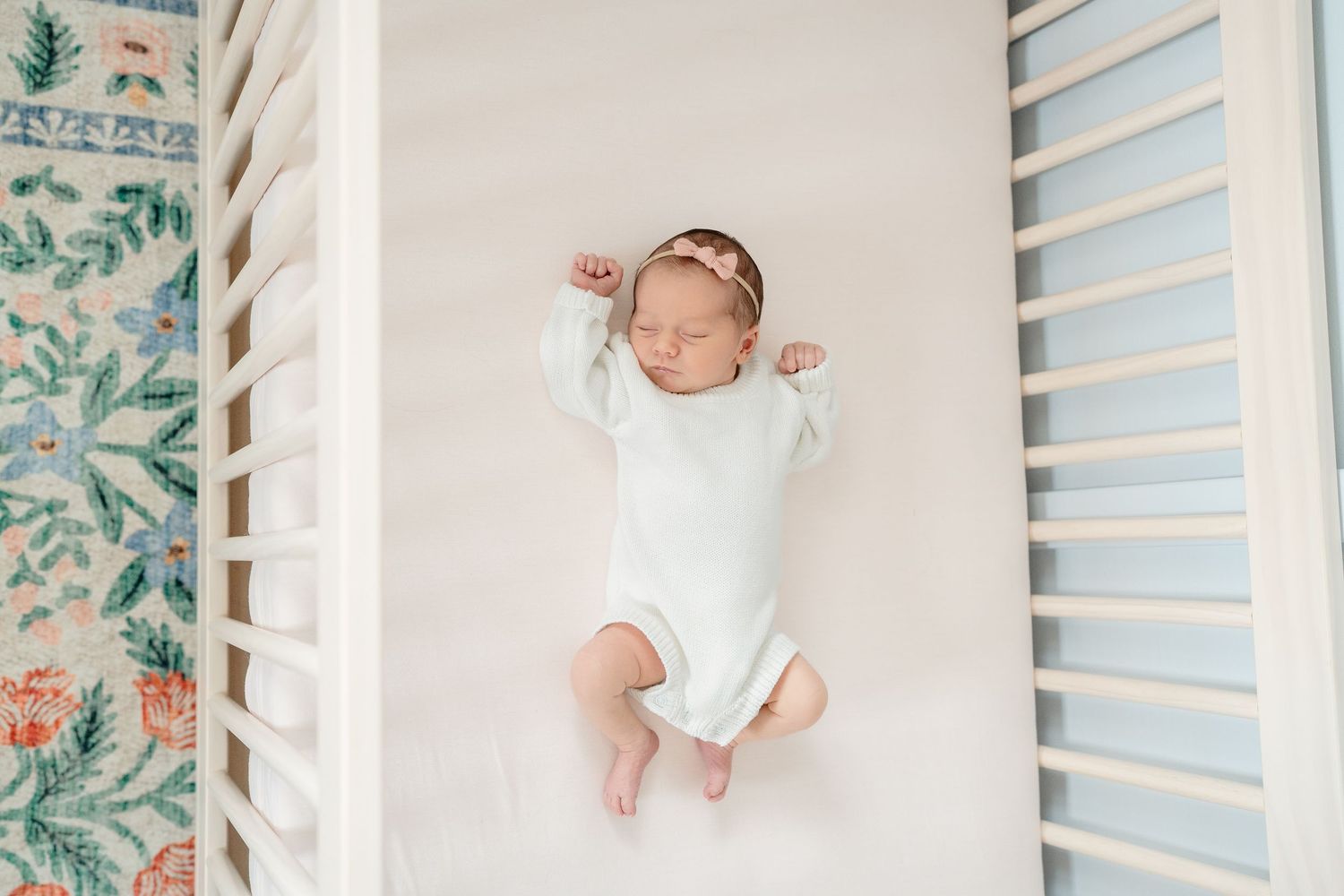 Newborn baby sleeping peacefully in a white crib wearing a white onesie and pink headband.