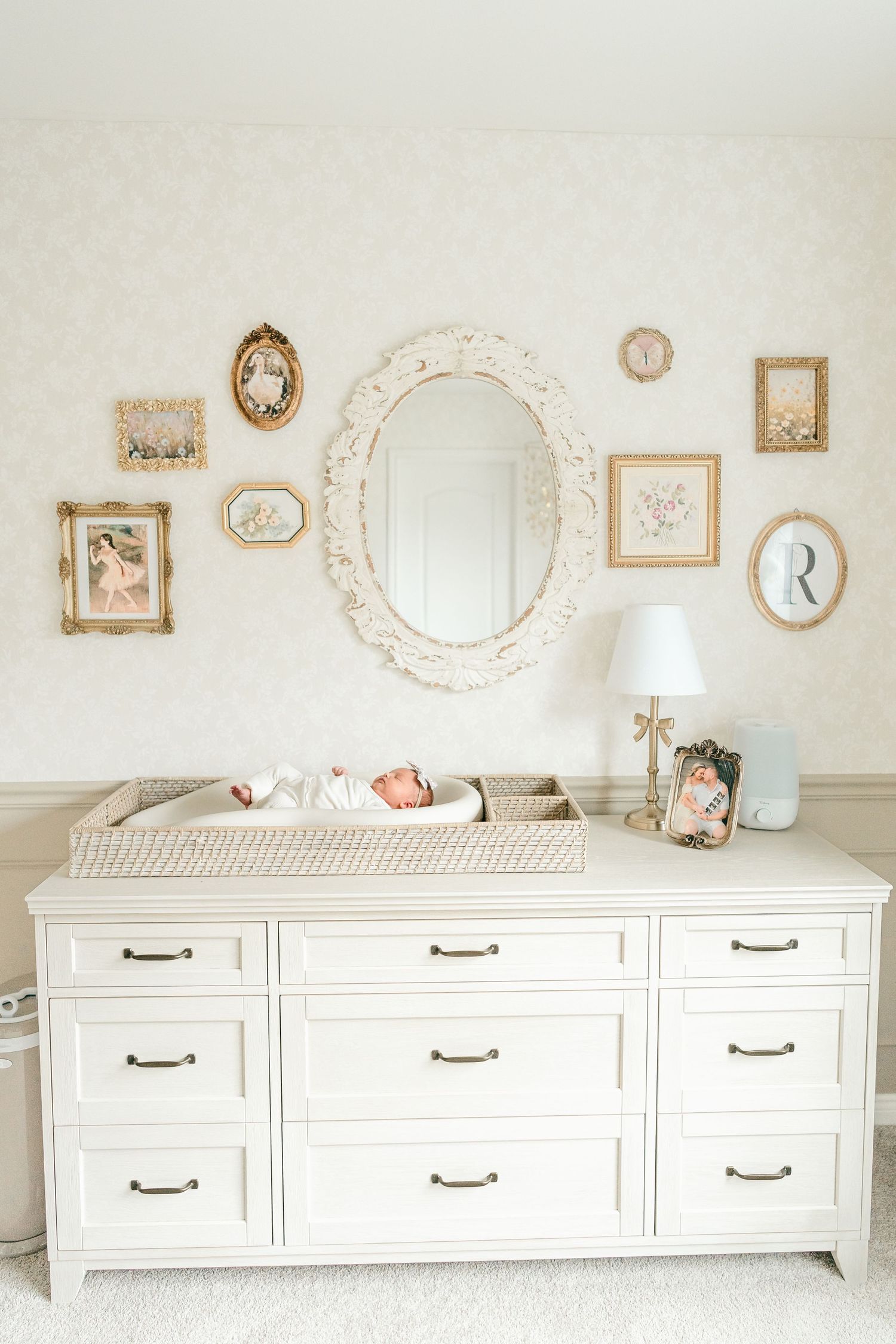 Newborn baby resting on a white changing dresser in an elegant nursery with vintage gold frames and ornate mirror.