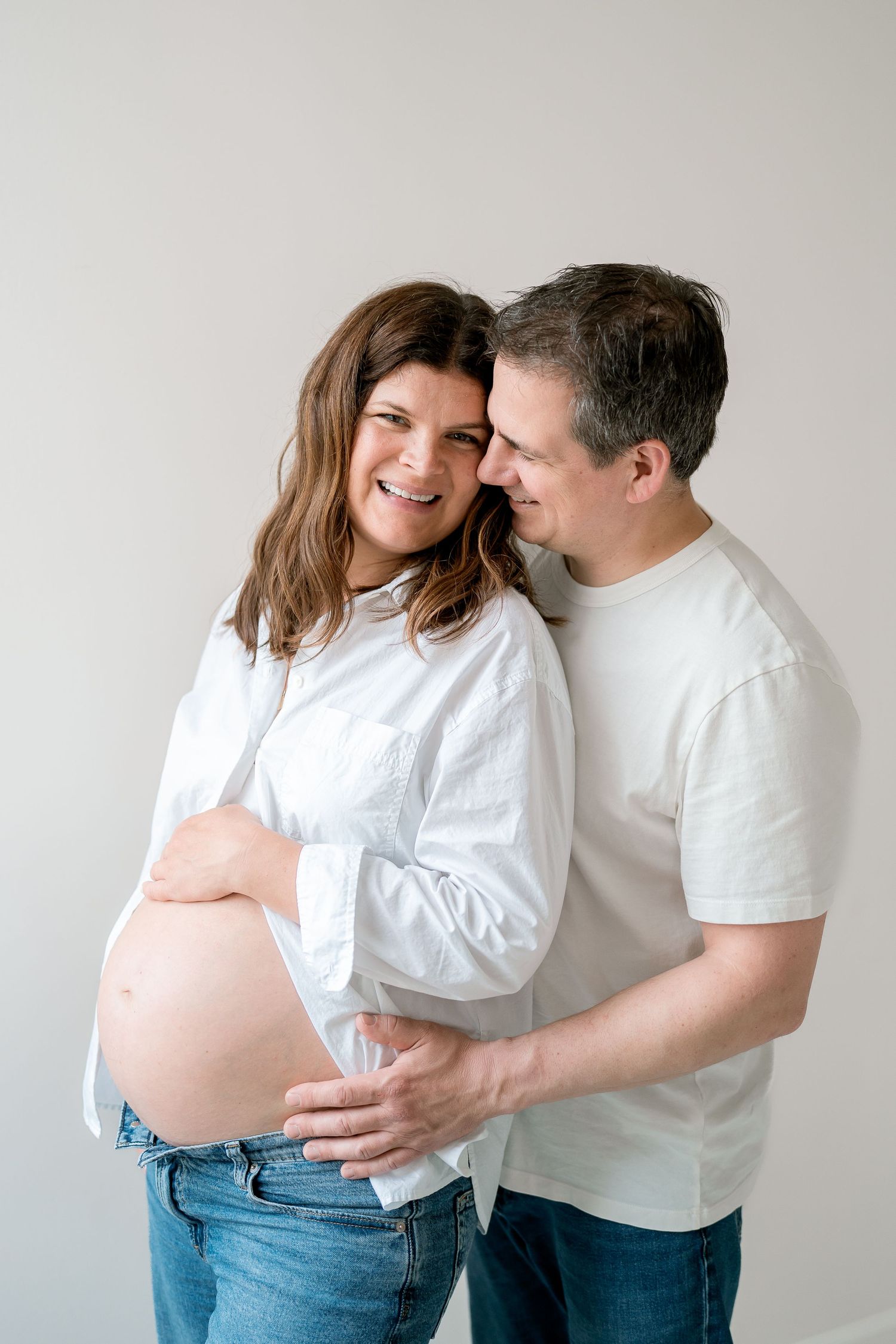 Smiling pregnant woman and her husband stand close together, both cradling her baby bump in a light studio.
