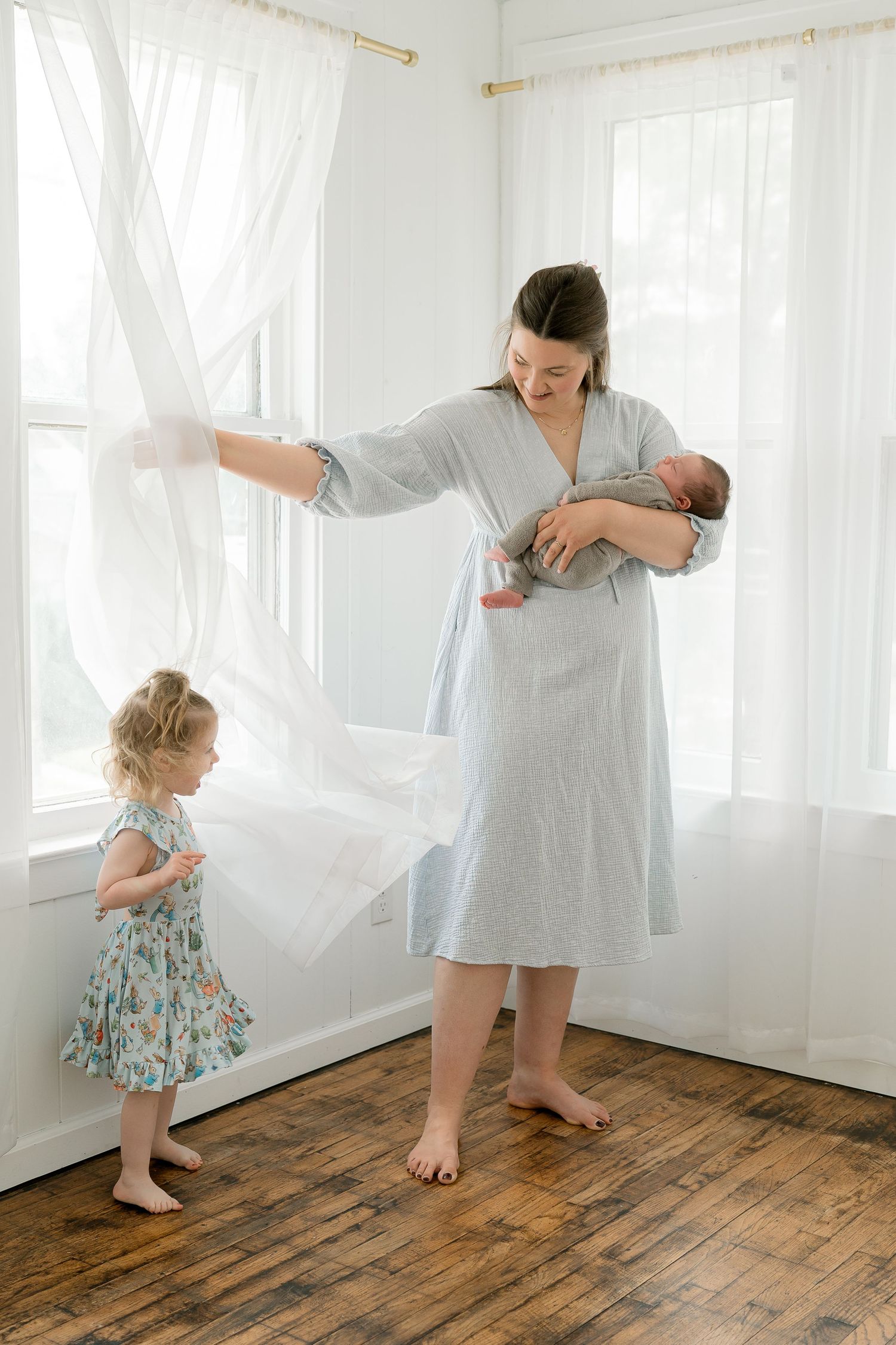 Mother in blue robe holds newborn while toddler girl plays with sheer curtains in bright white room.