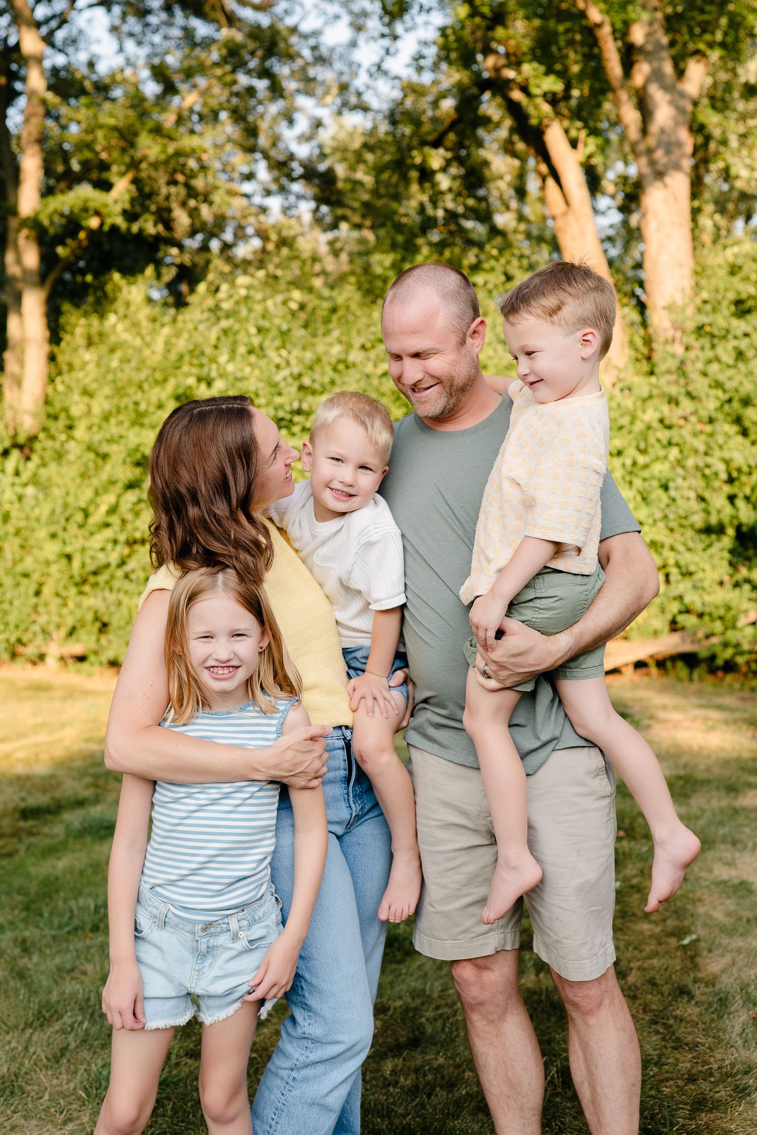 Happy family of five smiling outdoors, parents holding young children in a sunny green backyard setting.
