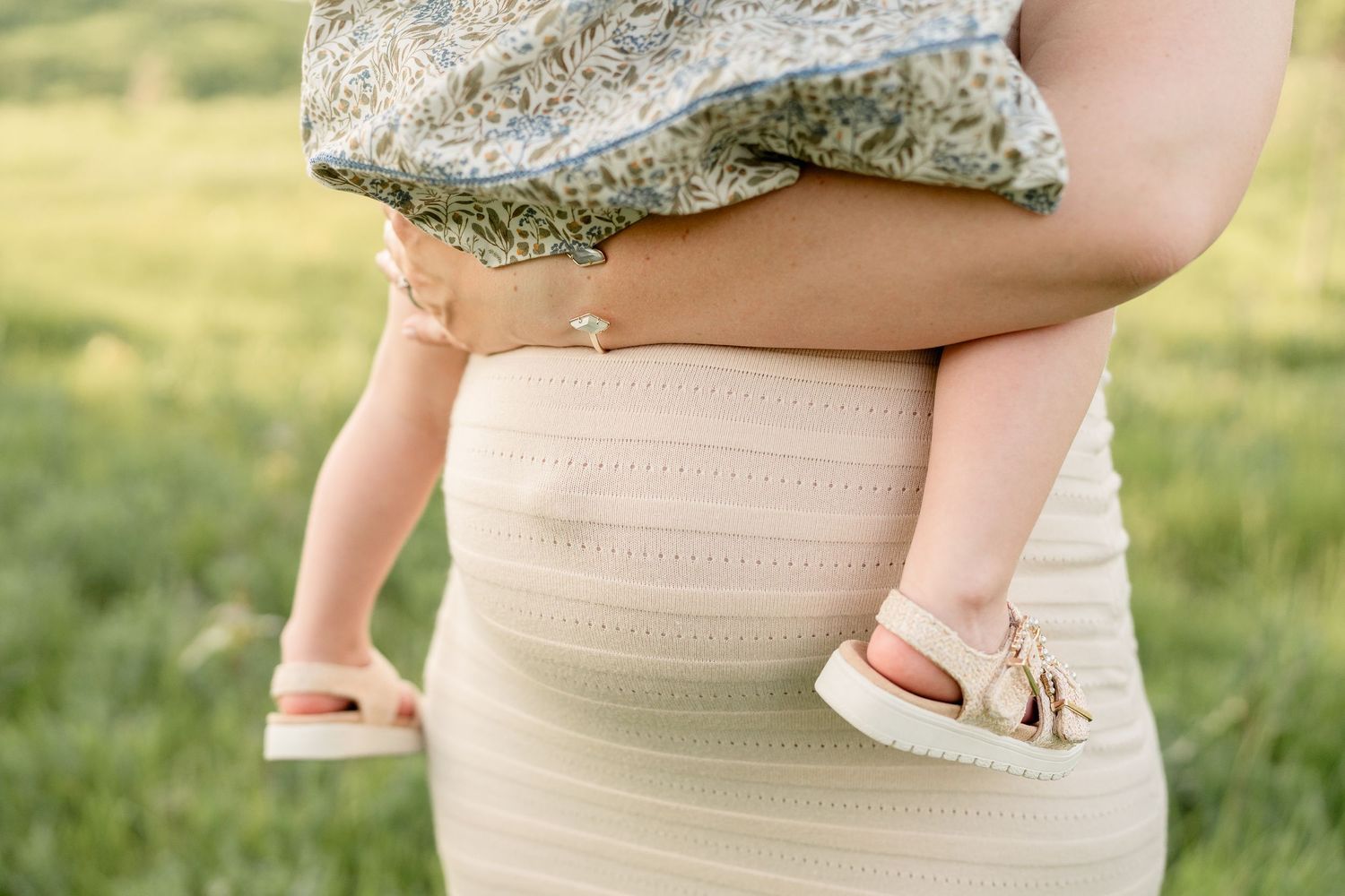 Close-up of pregnant woman in cream ribbed dress holding toddler in floral dress outdoors.