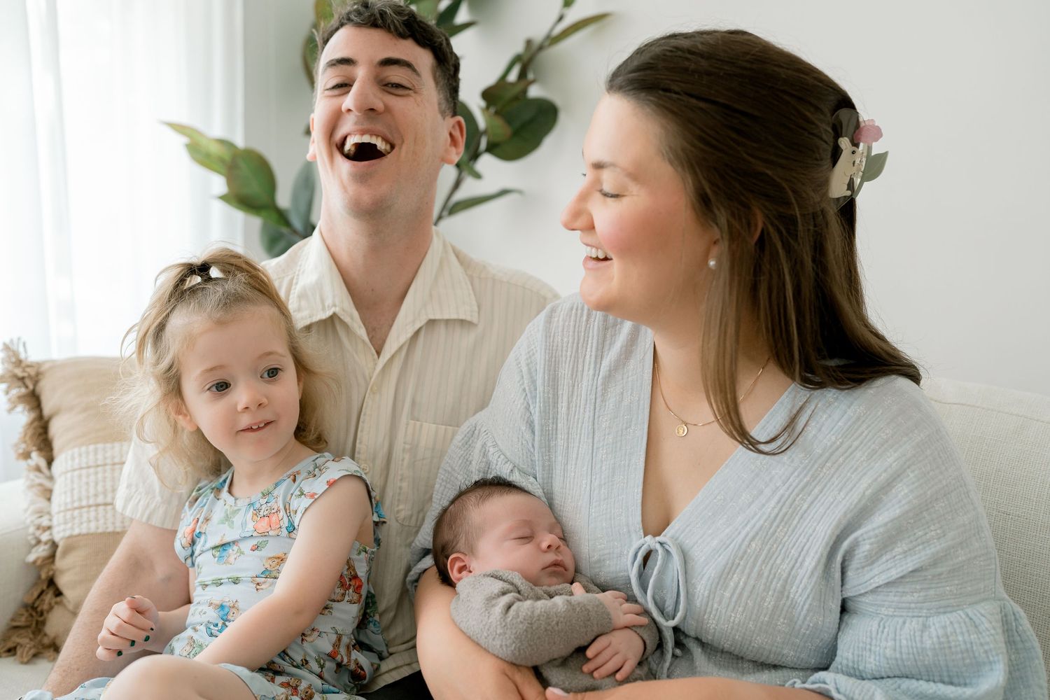 Joyful family with toddler girl and newborn baby laughing together on white couch.