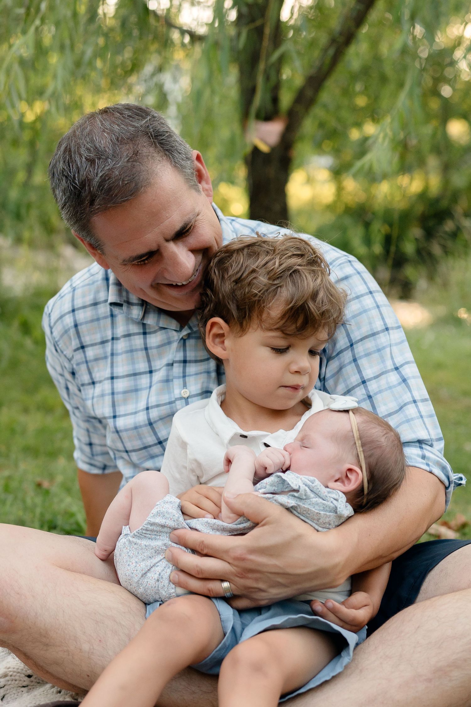 Father smiles holding newborn baby while young son gazes lovingly at sibling outdoors in green park setting.
