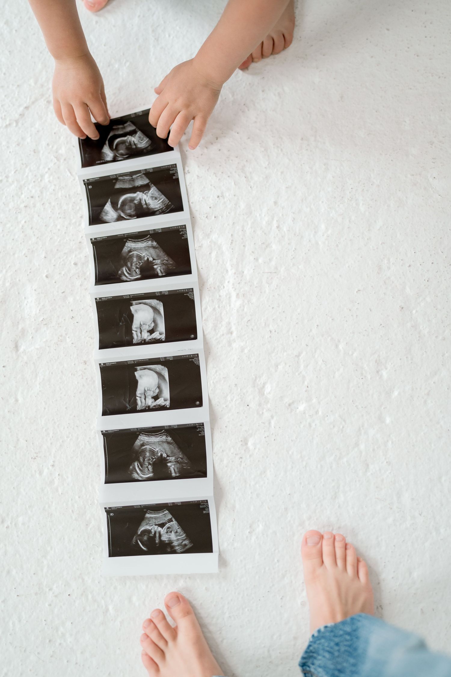 Child's hands arranging a row of baby ultrasound images on white floor during pregnancy announcement photo.