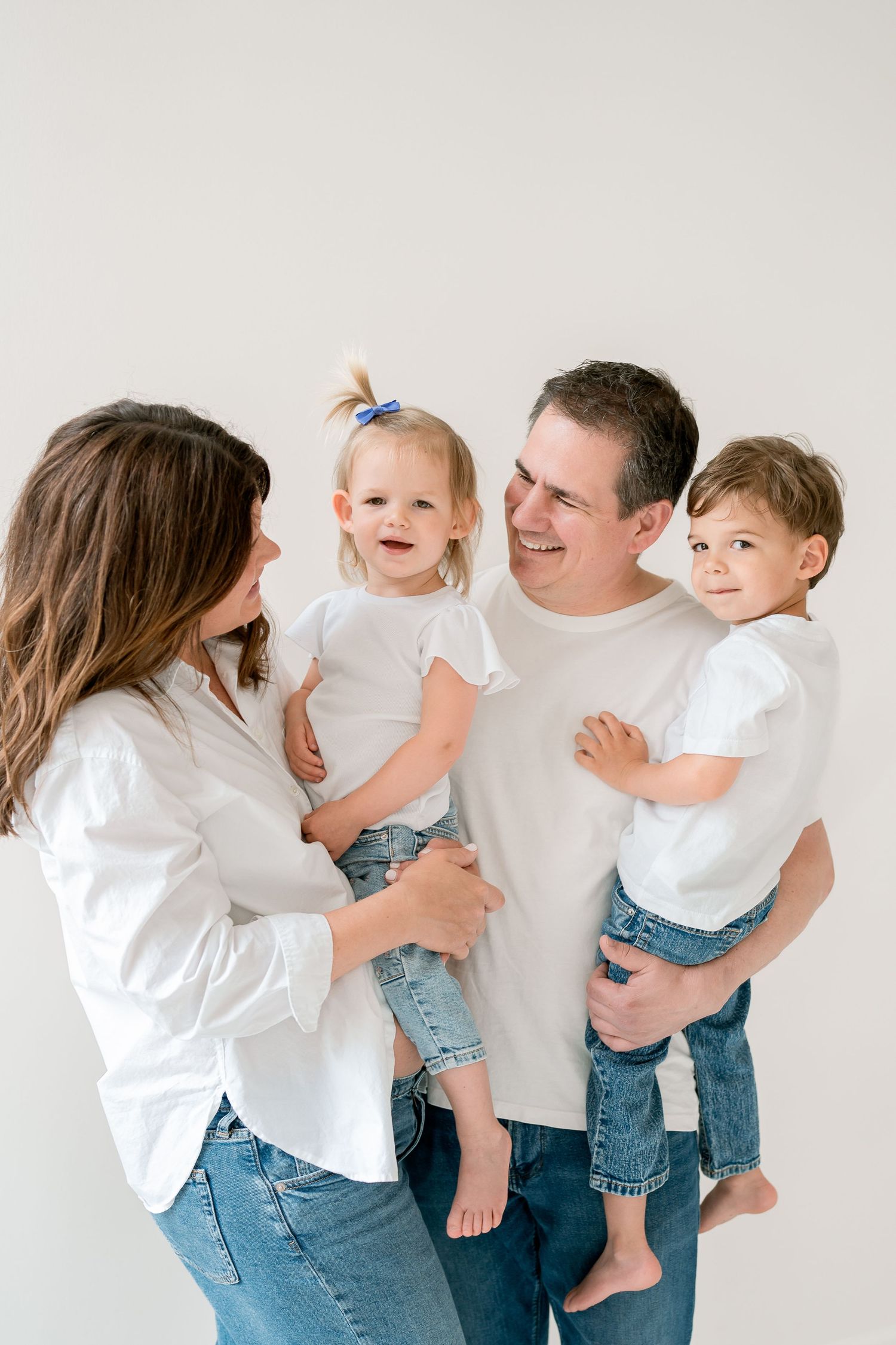 Happy family of four in matching white shirts and jeans, parents each holding a young child in a bright studio.