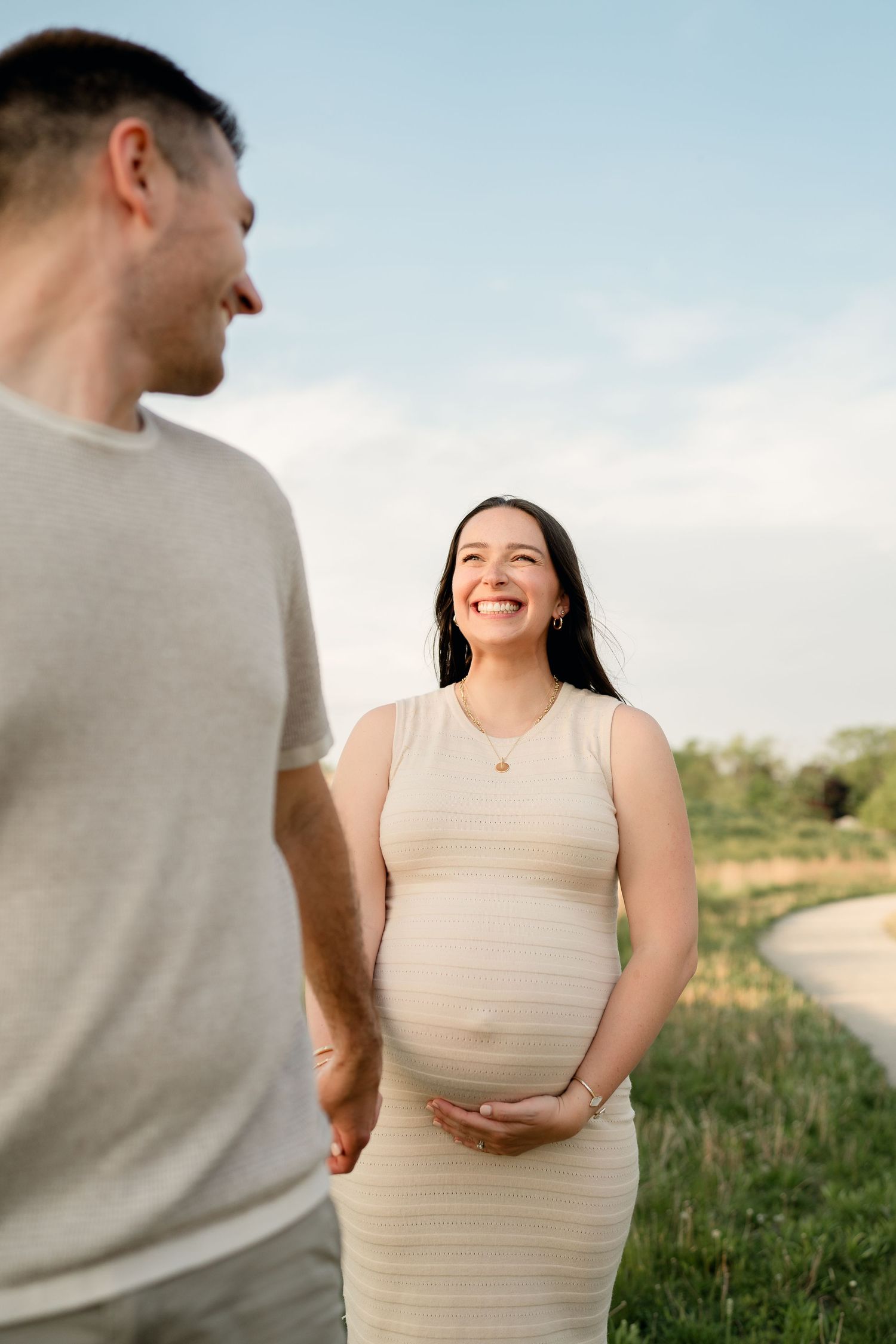 Smiling pregnant woman in cream dress holding belly while holding hands with partner outdoors.