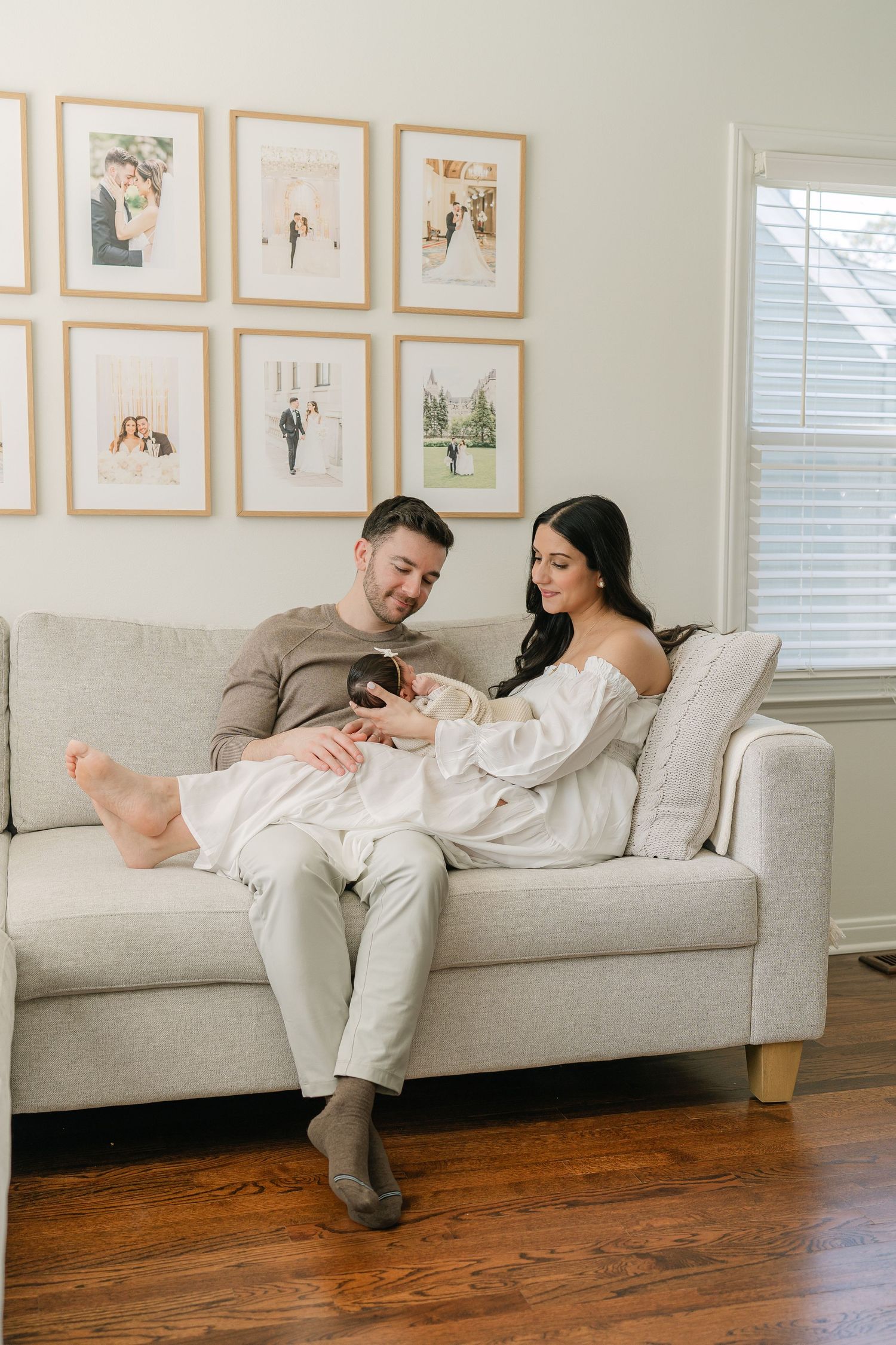 New parents relaxing on beige sofa with newborn baby, framed family photos displayed on wall behind them.