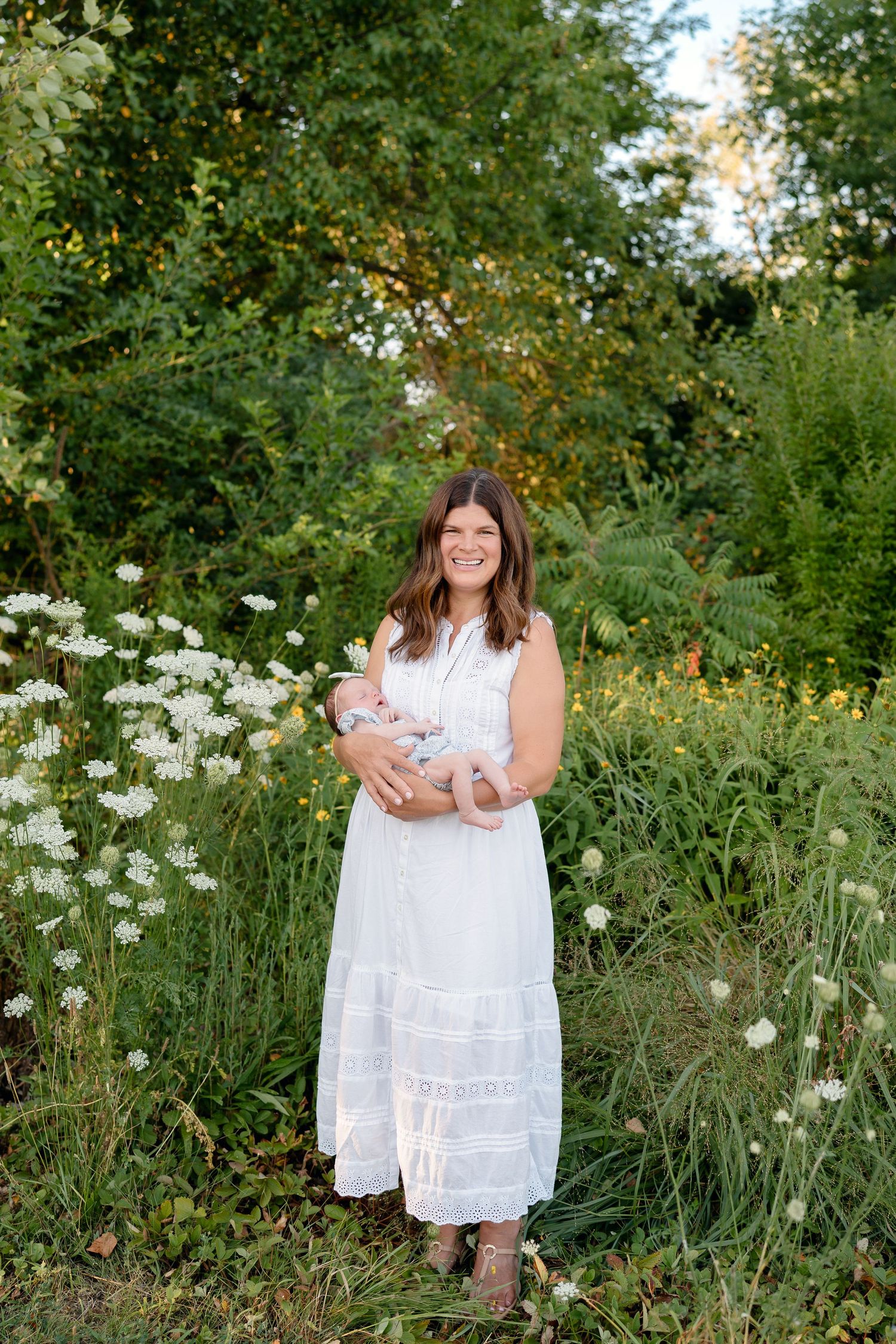 A smiling woman in a white dress holds a newborn baby while standing among wildflowers and lush green foliage outdoors.