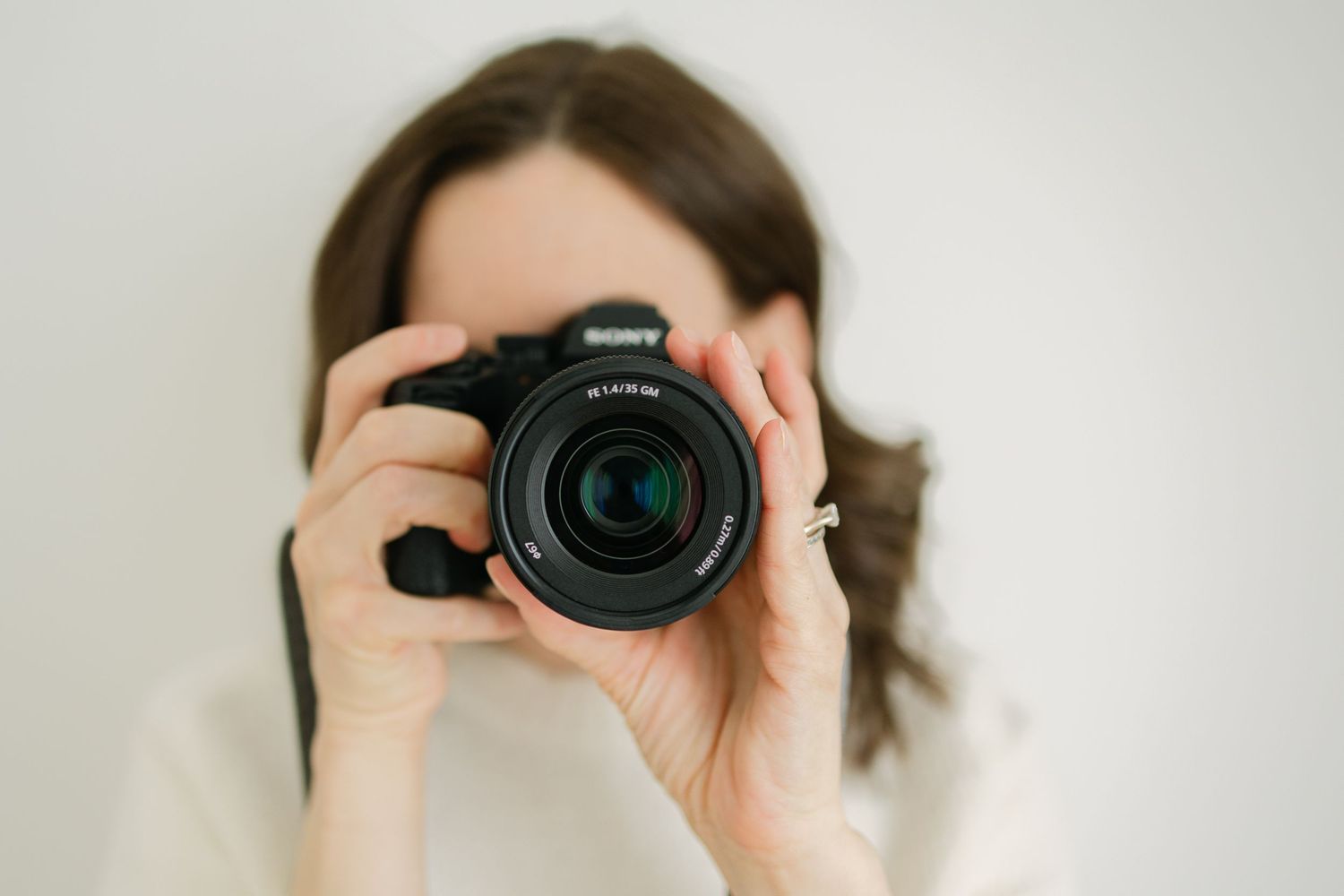 A female photographer holds a Sony DSLR camera up to her face against a light background.