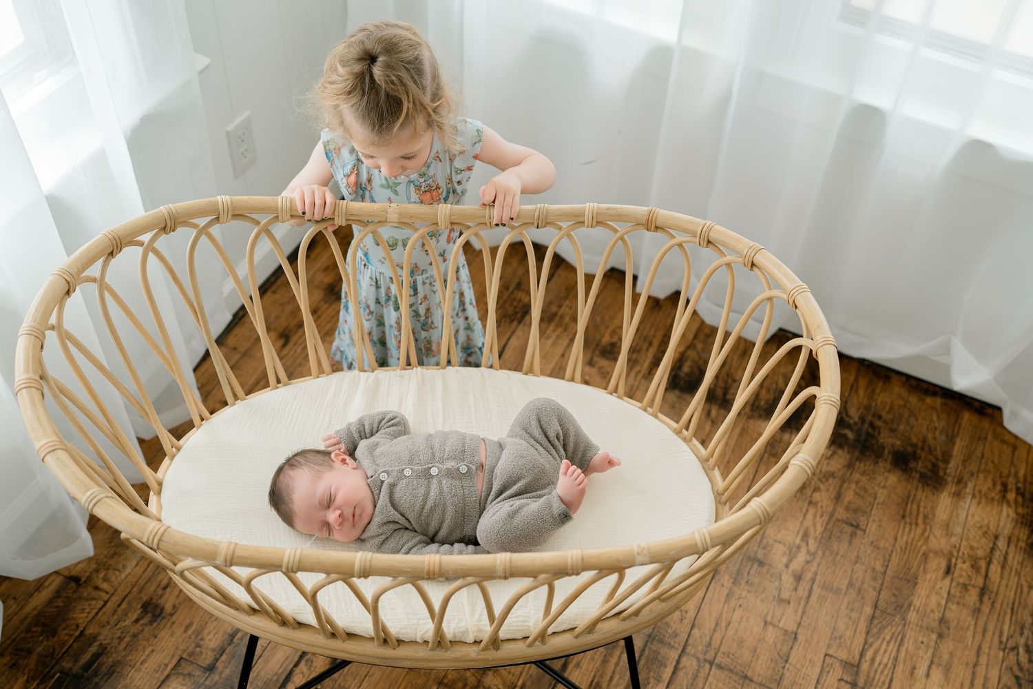 Toddler girl in floral dress looking at sleeping newborn baby in a wicker bassinet on hardwood floors.