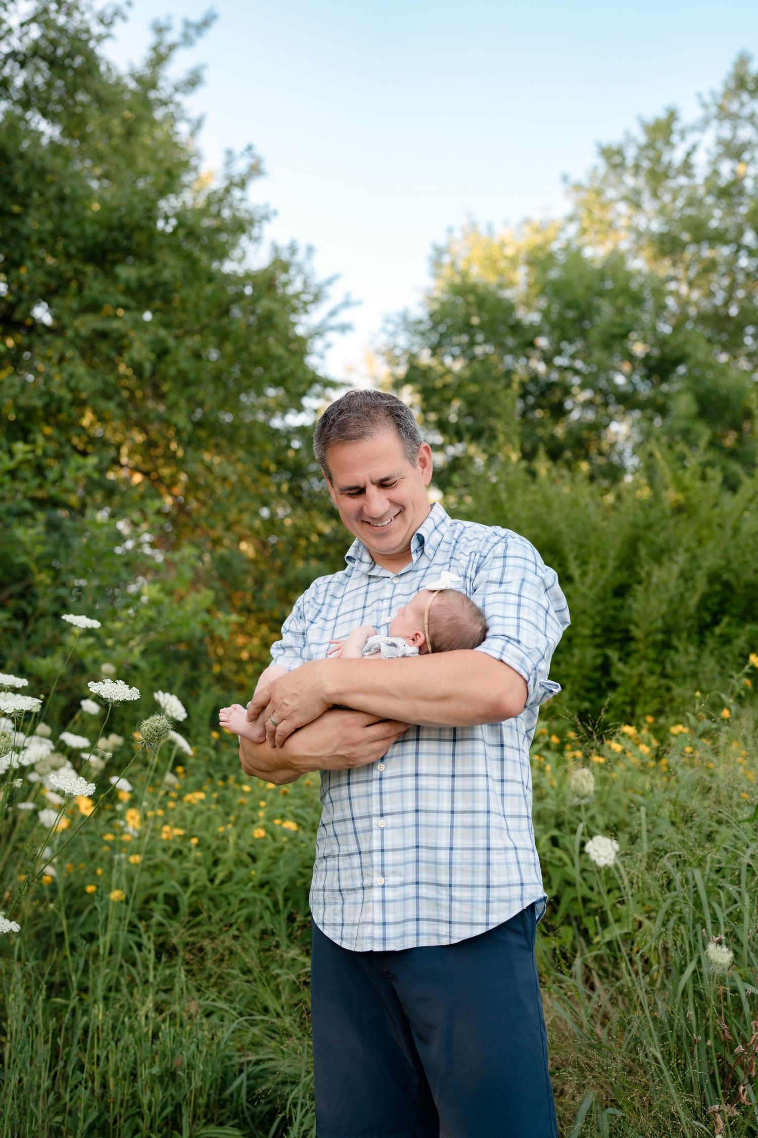 A smiling man holds a newborn baby outdoors, surrounded by wildflowers and lush green trees.