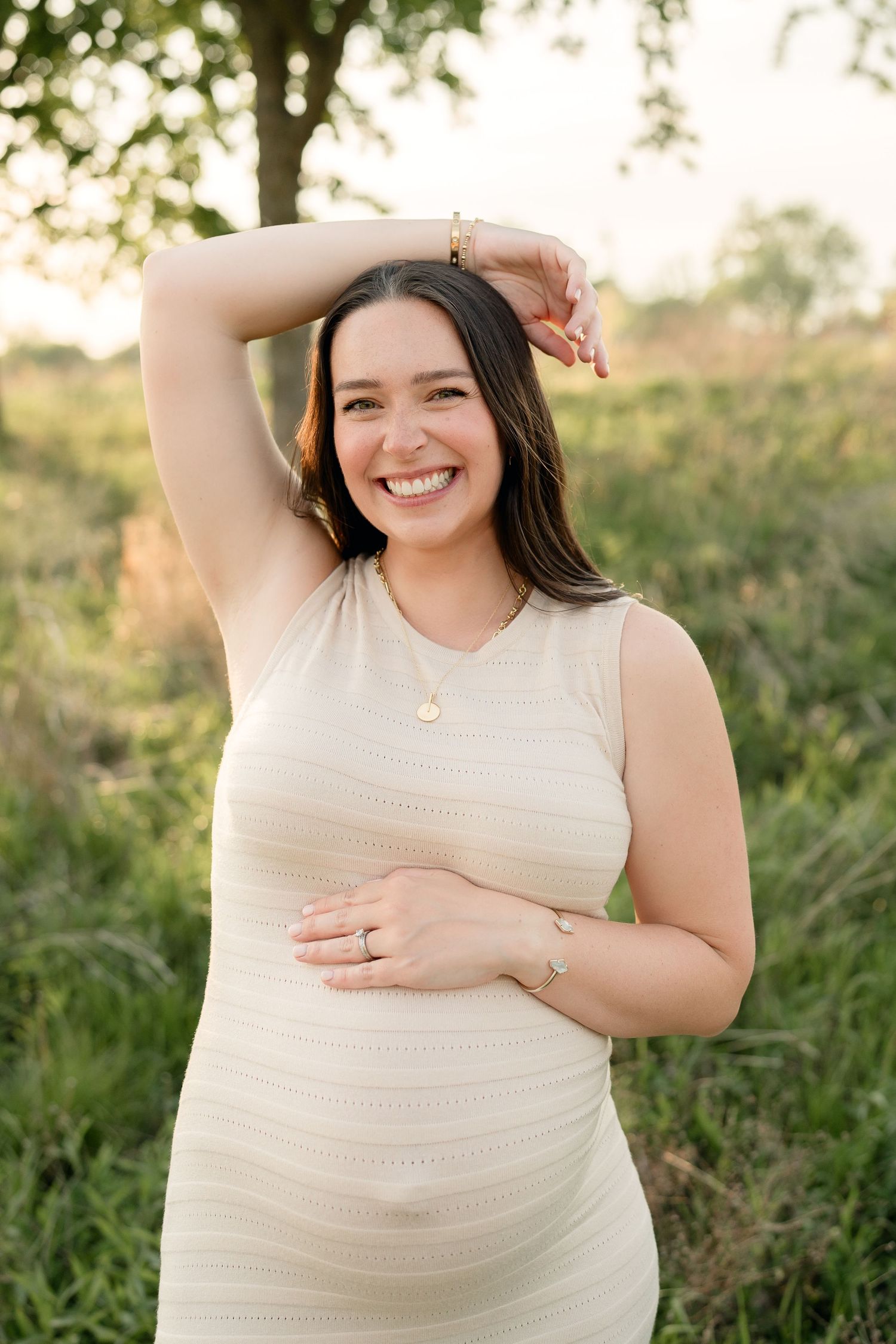 Joyful pregnant woman in cream dress posing with arm raised in lush green field.