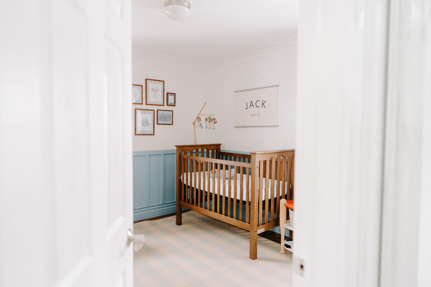 Wooden baby crib in a neutral nursery with blue wainscoting and Jack name sign.