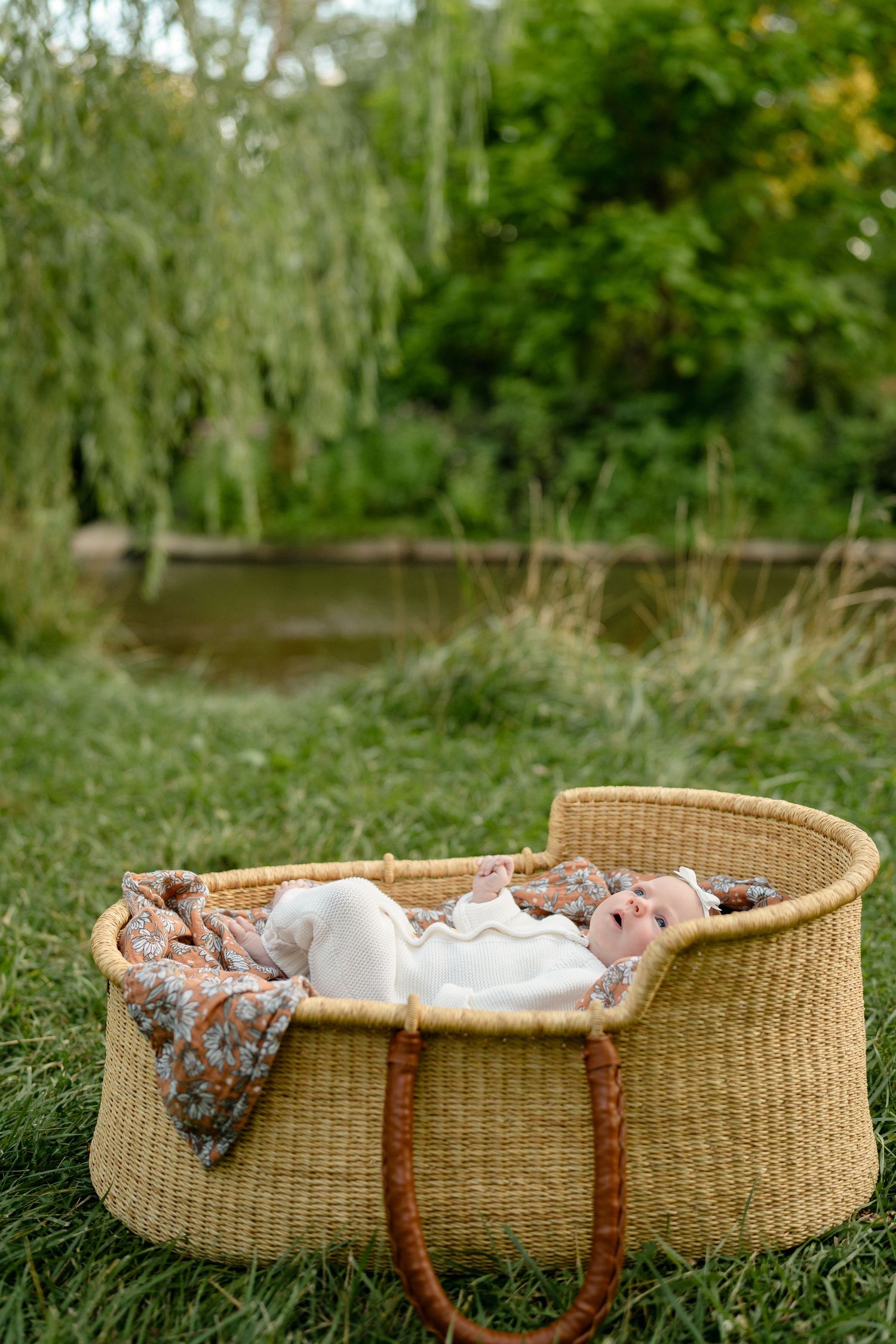 Newborn baby lying in a wicker Moses basket on green grass outdoors near a pond.