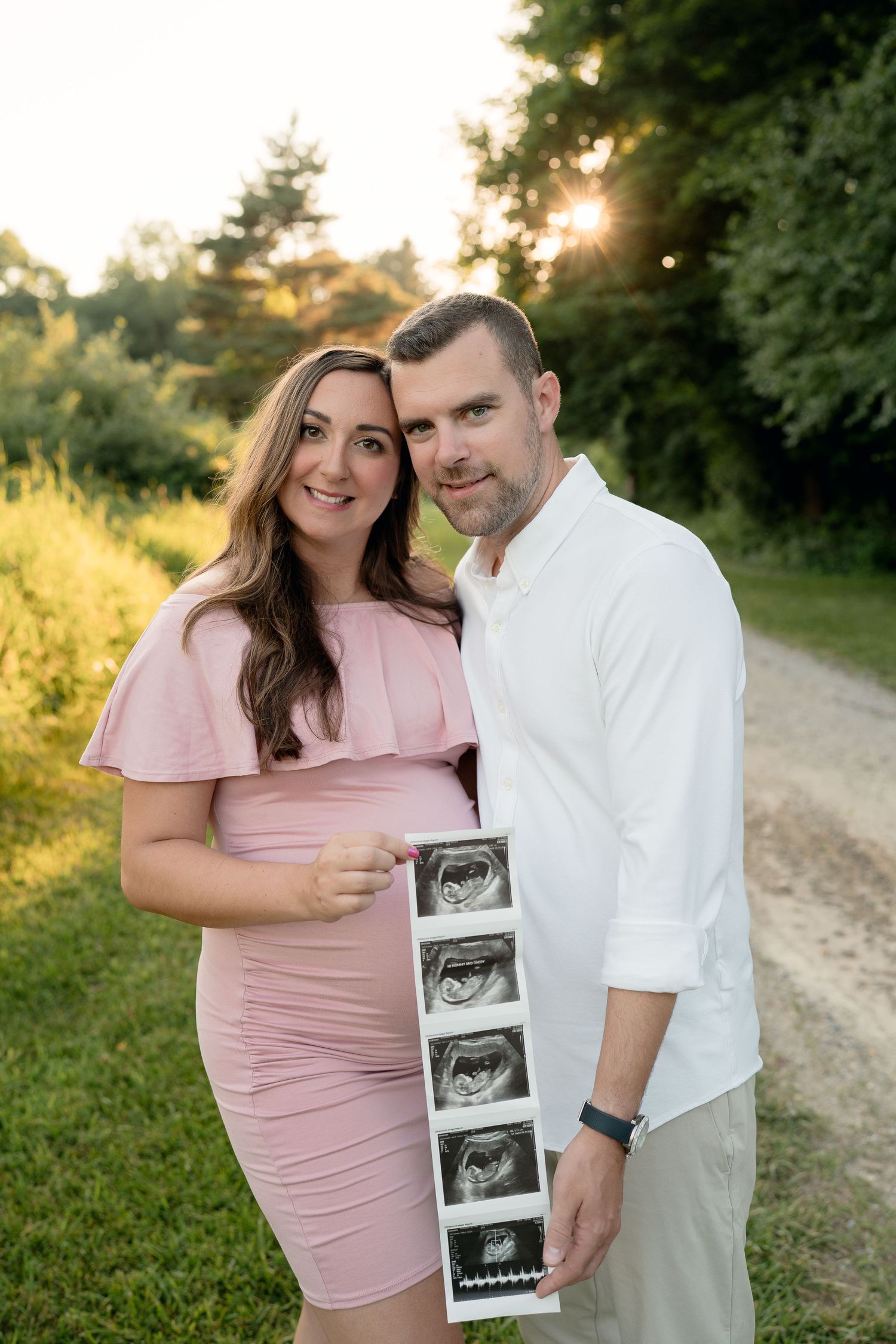 Smiling expecting couple holding ultrasound photos on a sunlit path during a golden hour outdoor maternity session.