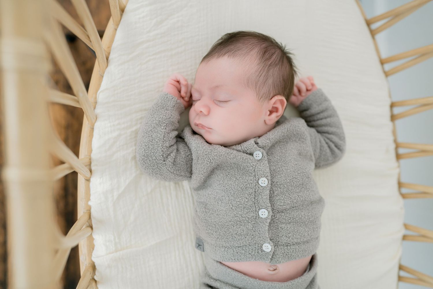 A peaceful newborn baby sleeping in a white bassinet wearing a soft gray knit cardigan.