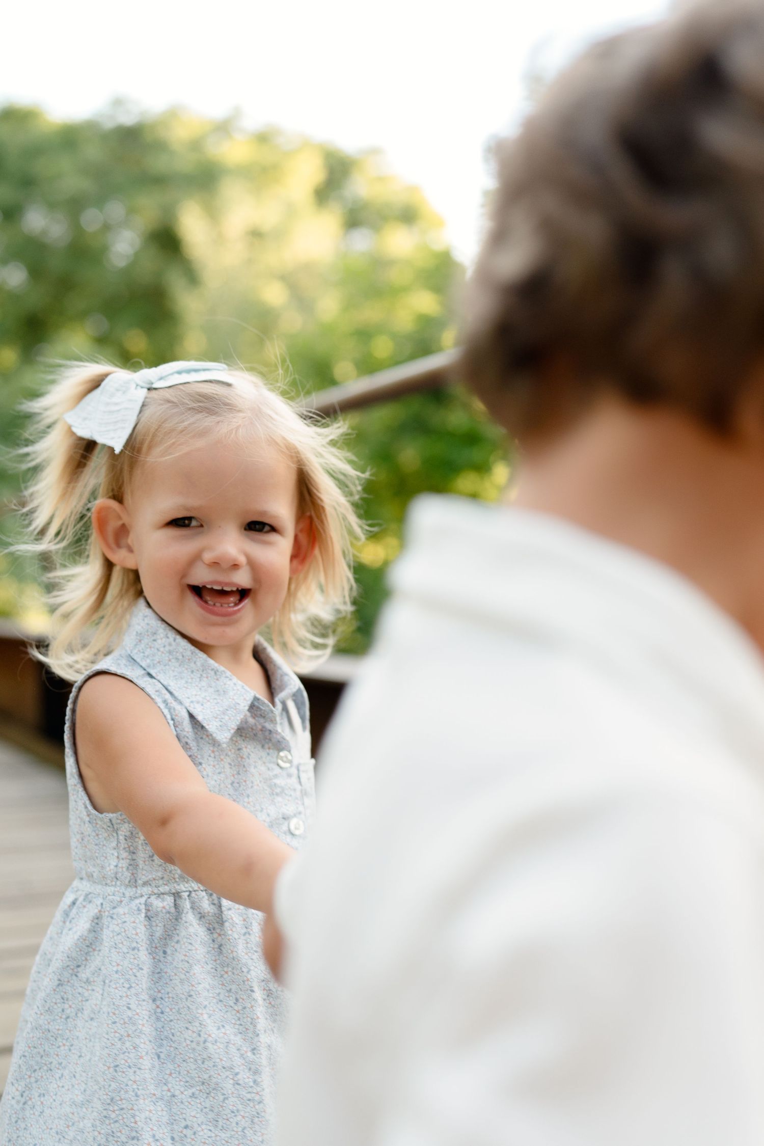 Happy toddler girl with blonde pigtails in blue dress laughing and holding hands with adult outdoors.