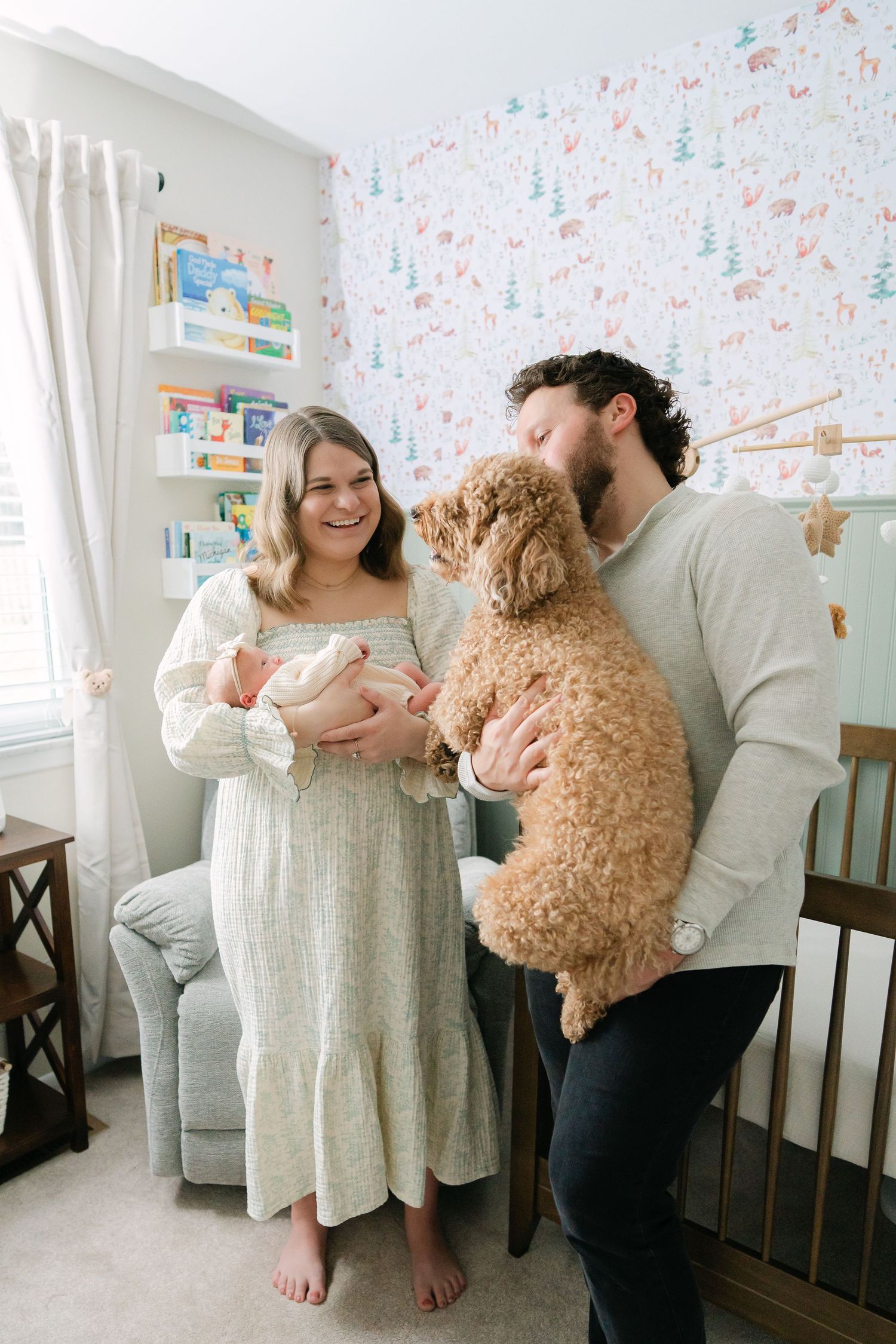 Happy parents holding newborn baby and golden doodle dog in a charming woodland wallpaper nursery.