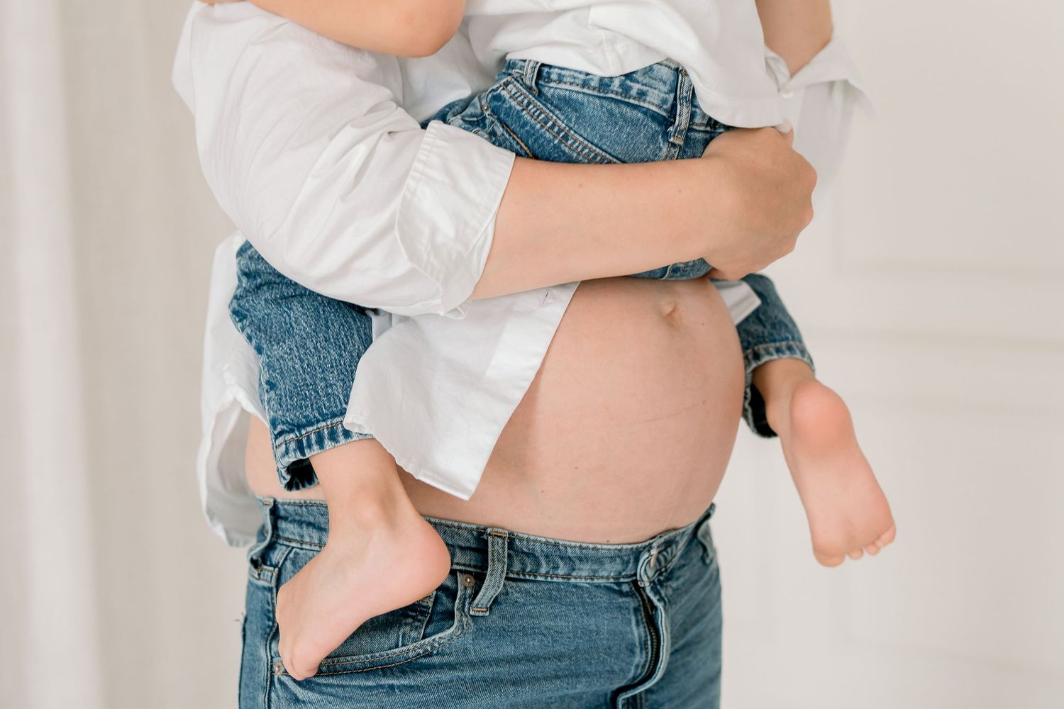 Close-up of a pregnant woman in a white shirt and jeans cradling a young child against her baby bump.