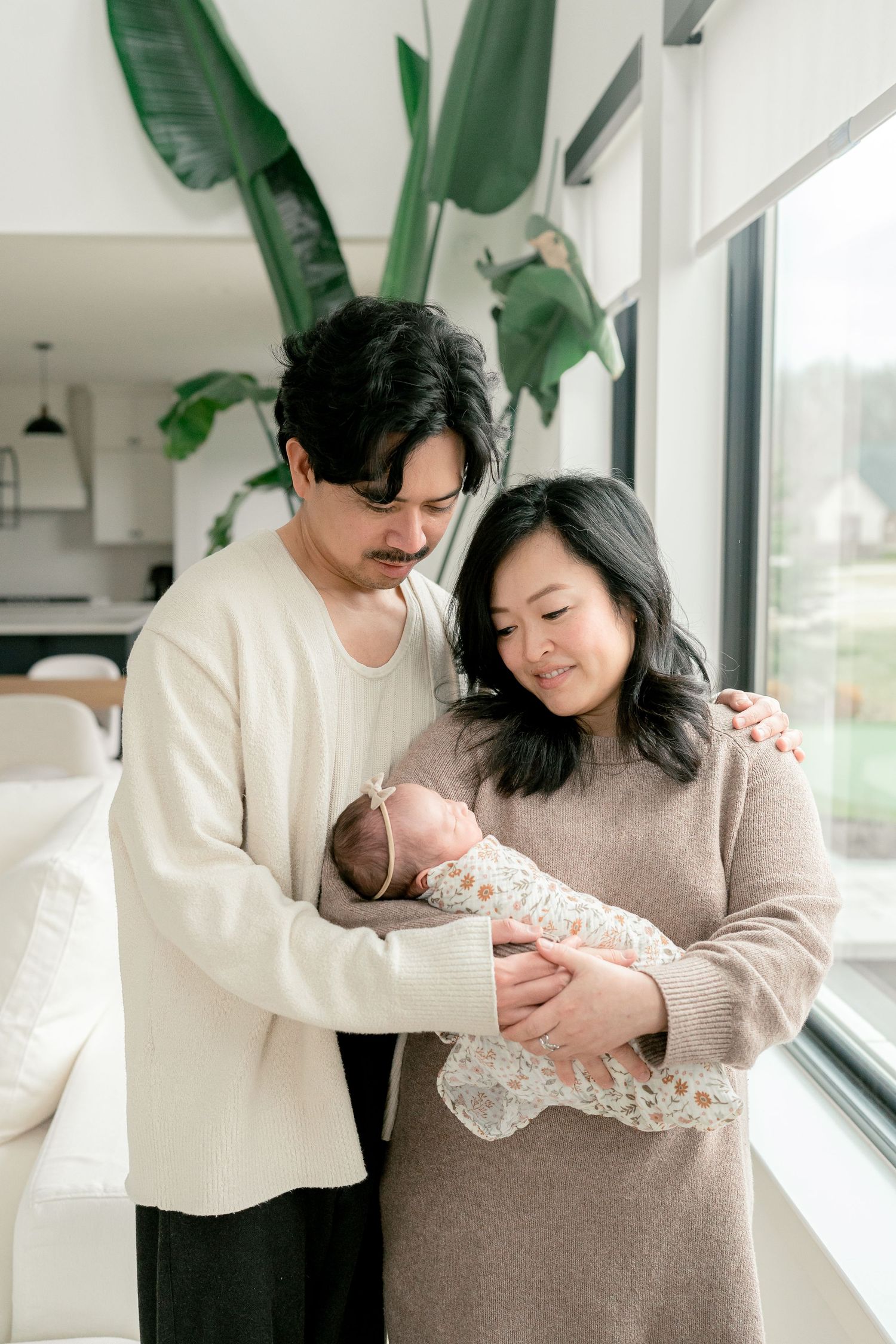 Couple gazes lovingly at swaddled newborn baby standing by floor-to-ceiling windows in bright modern living room in Oakland County, MI.