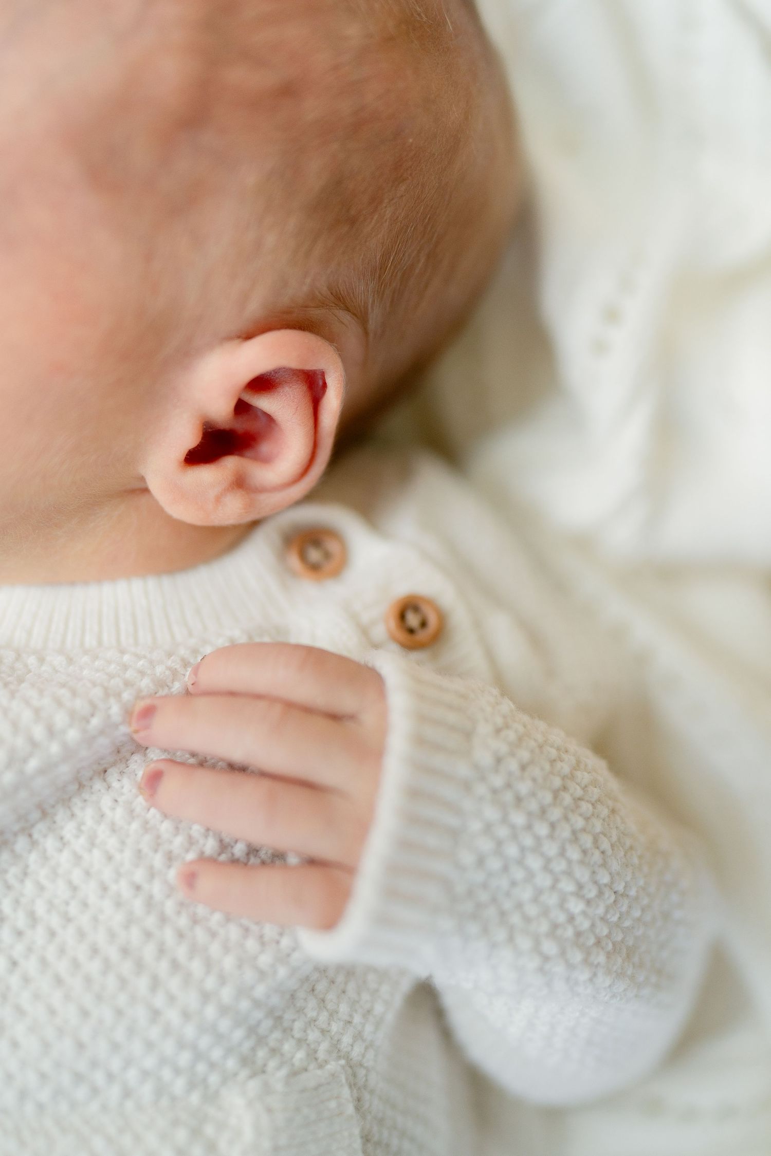 Close-up of a newborn babys tiny hand and ear in a cream knit sweater with wooden buttons.