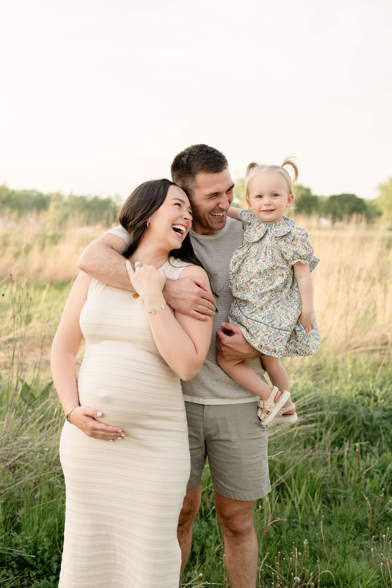 Happy pregnant family with father holding toddler girl laughing together in golden field.