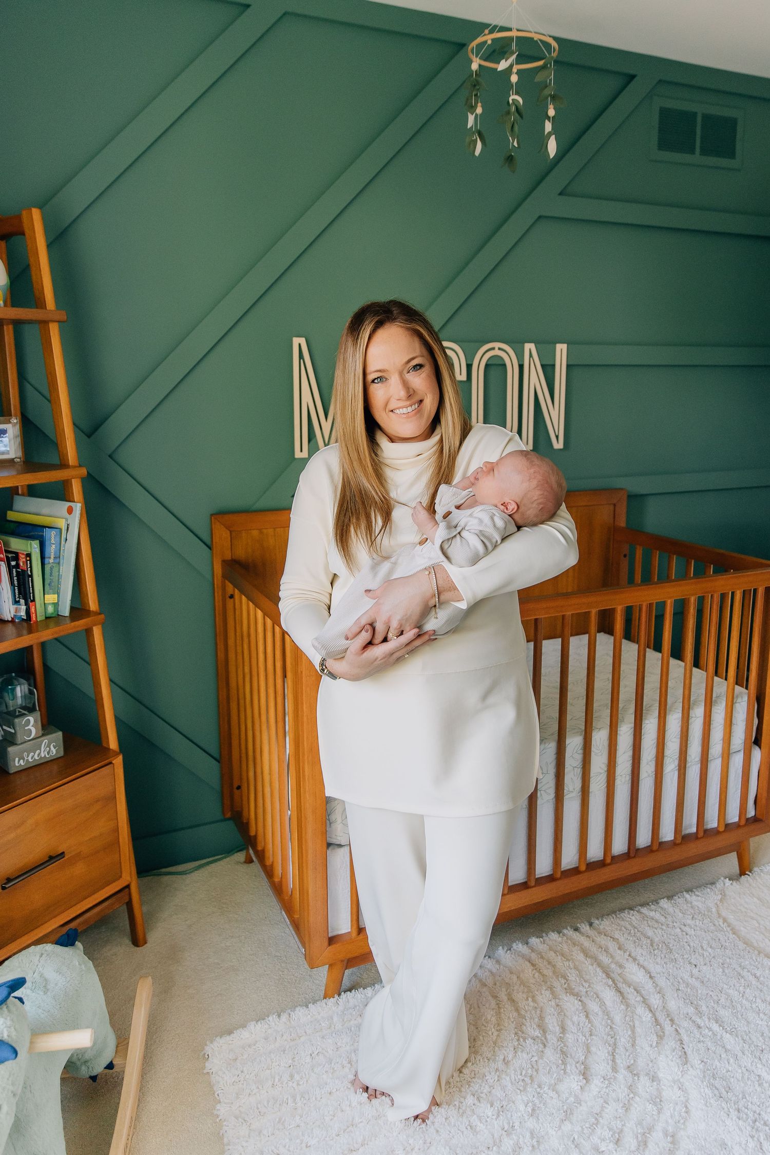 Smiling blonde mother in cream outfit holds newborn in green nursery with Mason name sign.