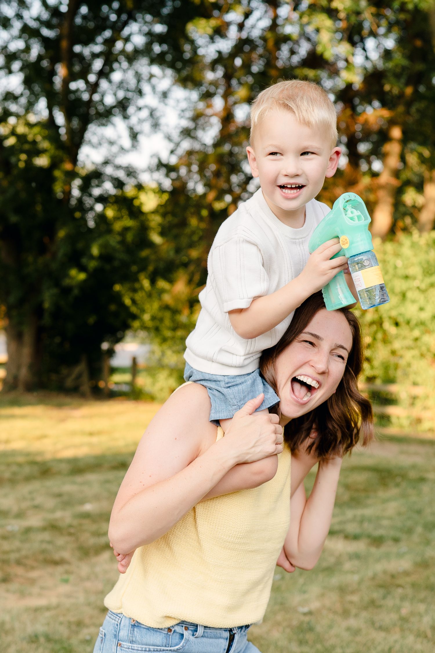 A joyful mother carries her laughing toddler son on her shoulders in a sunny park, both smiling brightly.
