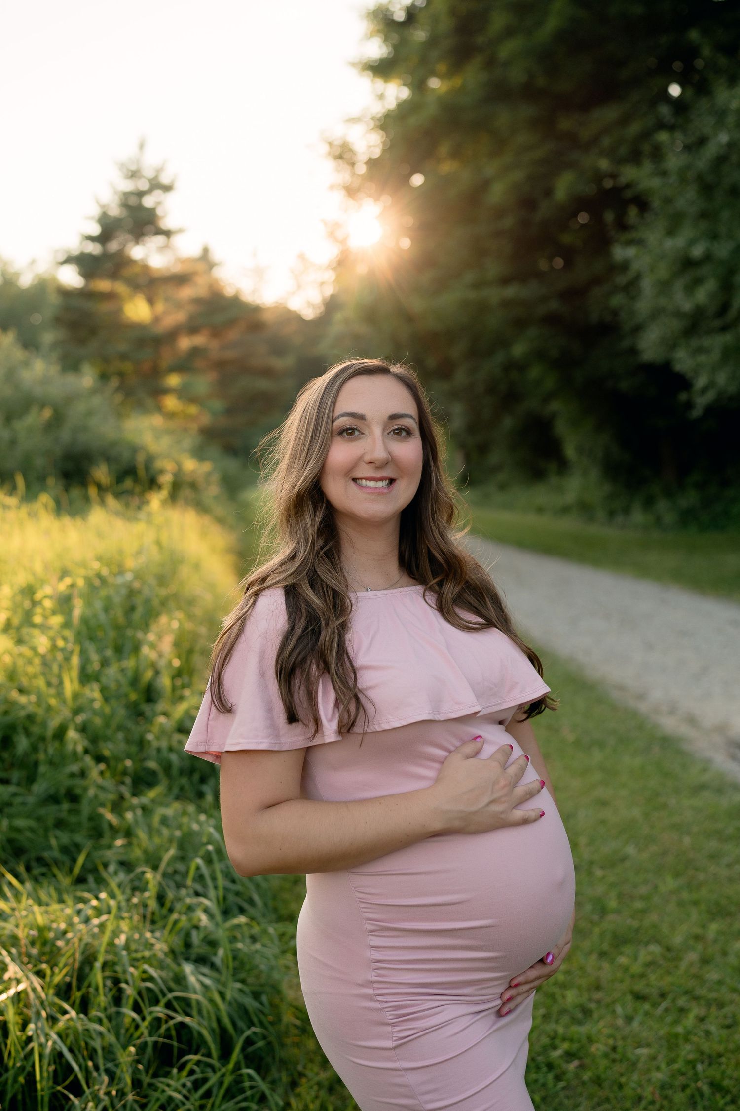 Smiling pregnant woman in pink dress holds baby bump outdoors at golden hour.