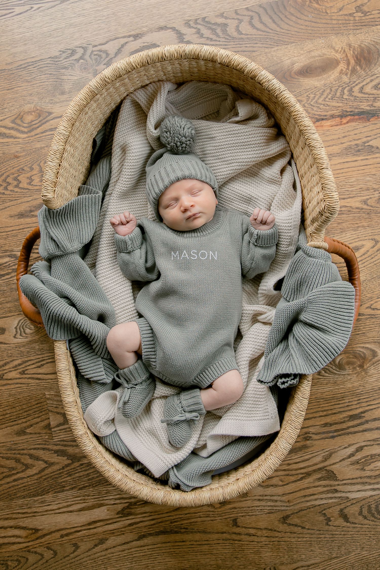Sleeping newborn baby named Mason in sage green knit outfit and pom hat rests in a woven Moses basket.