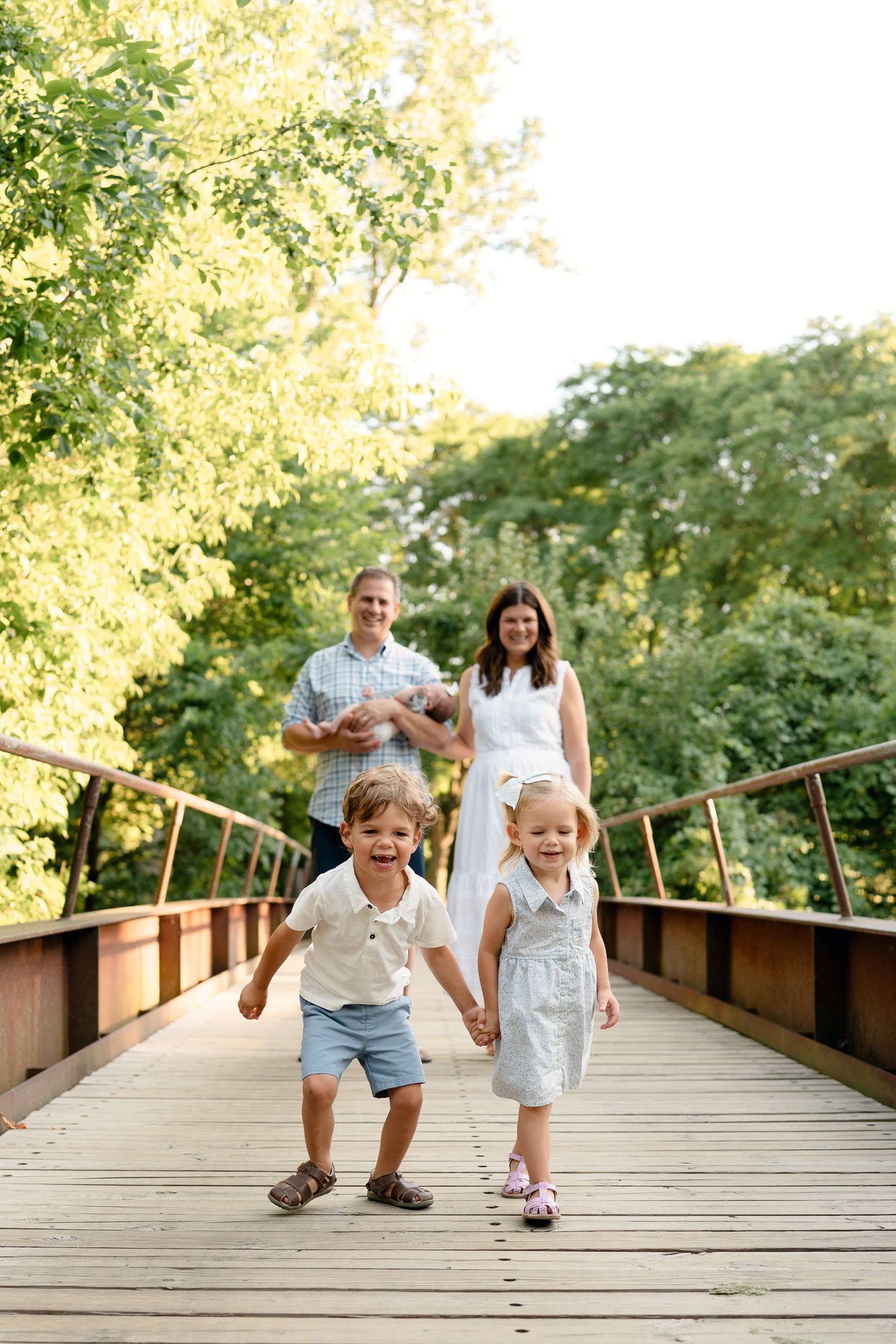 Family of four with newborn poses on wooden bridge surrounded by lush green trees on sunny day.