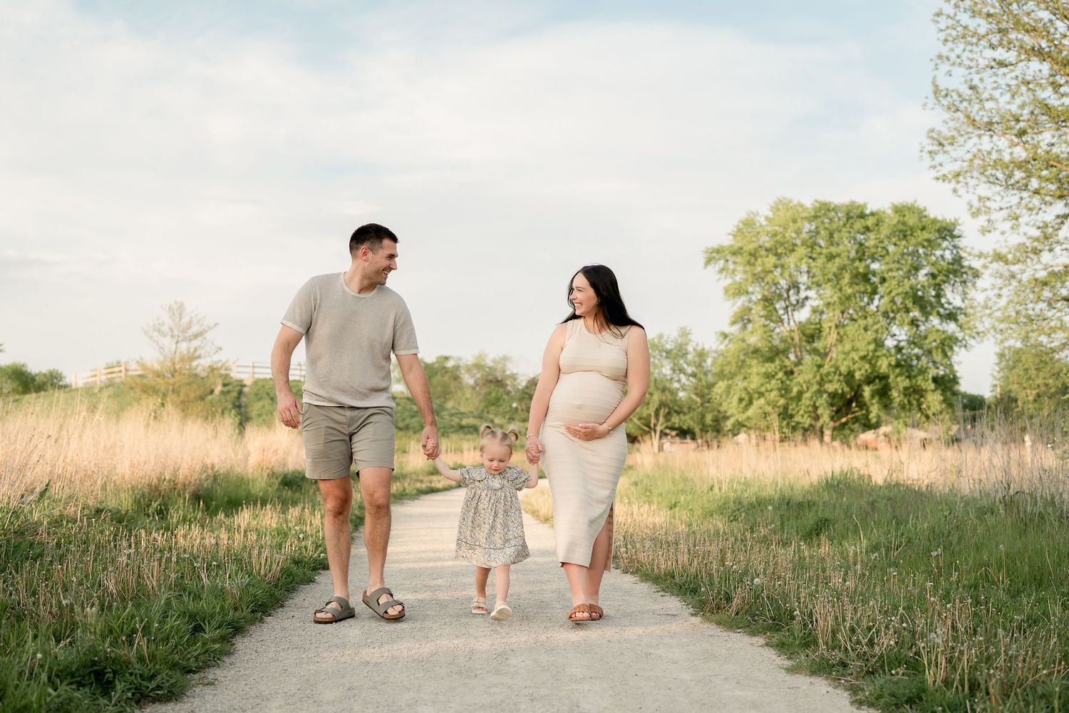 Happy pregnant family walking together on path through green meadow with toddler daughter.
