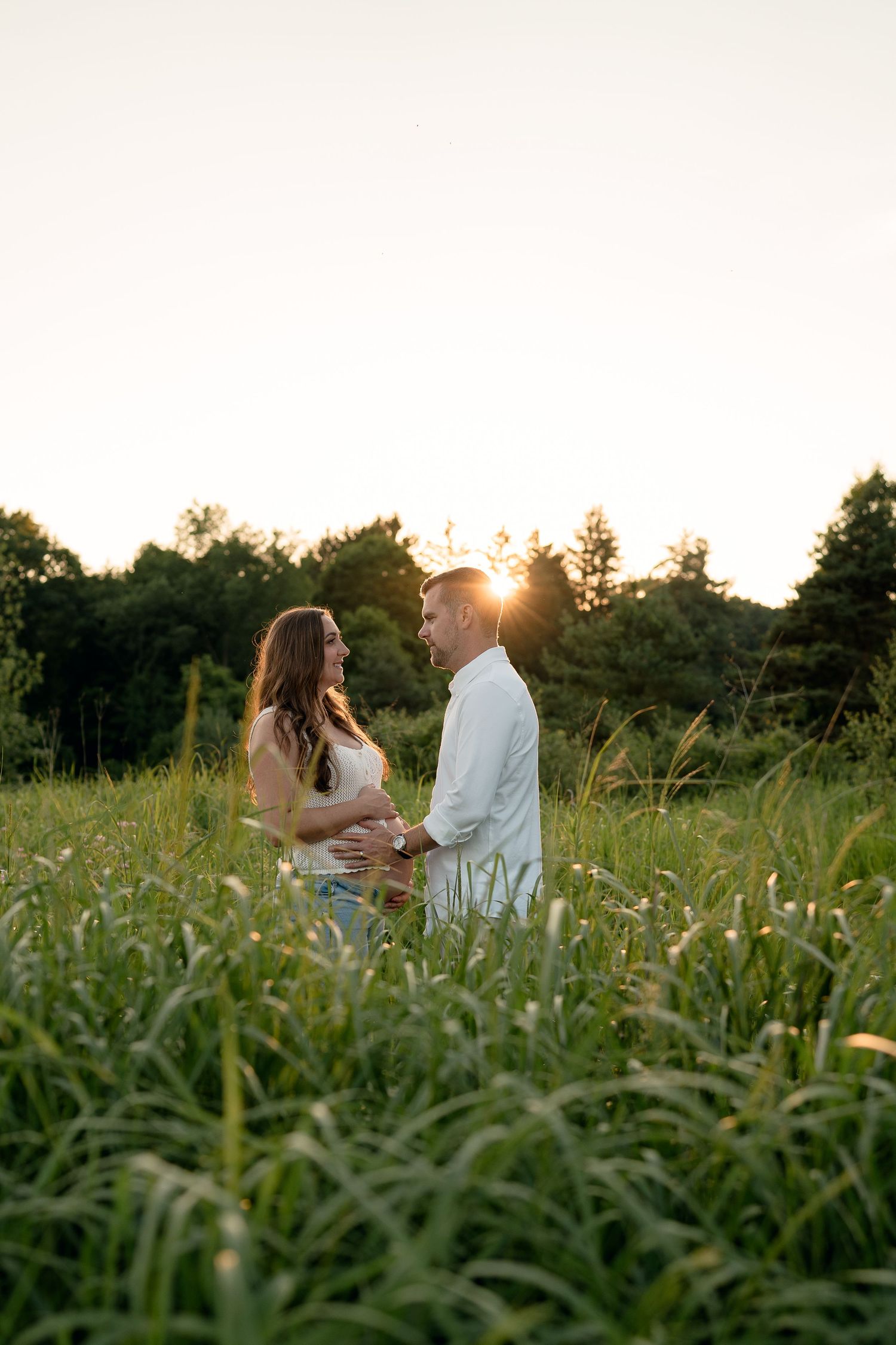 Pregnant couple facing each other holding hands in tall grass at golden sunset.