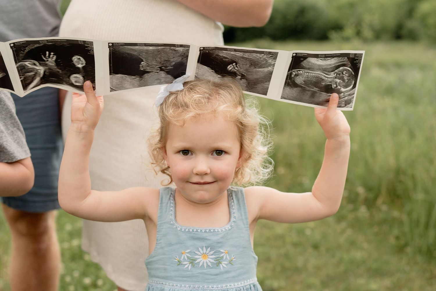 Curly-haired toddler girl in blue dress holds up ultrasound photos outdoors, pregnancy announcement concept.