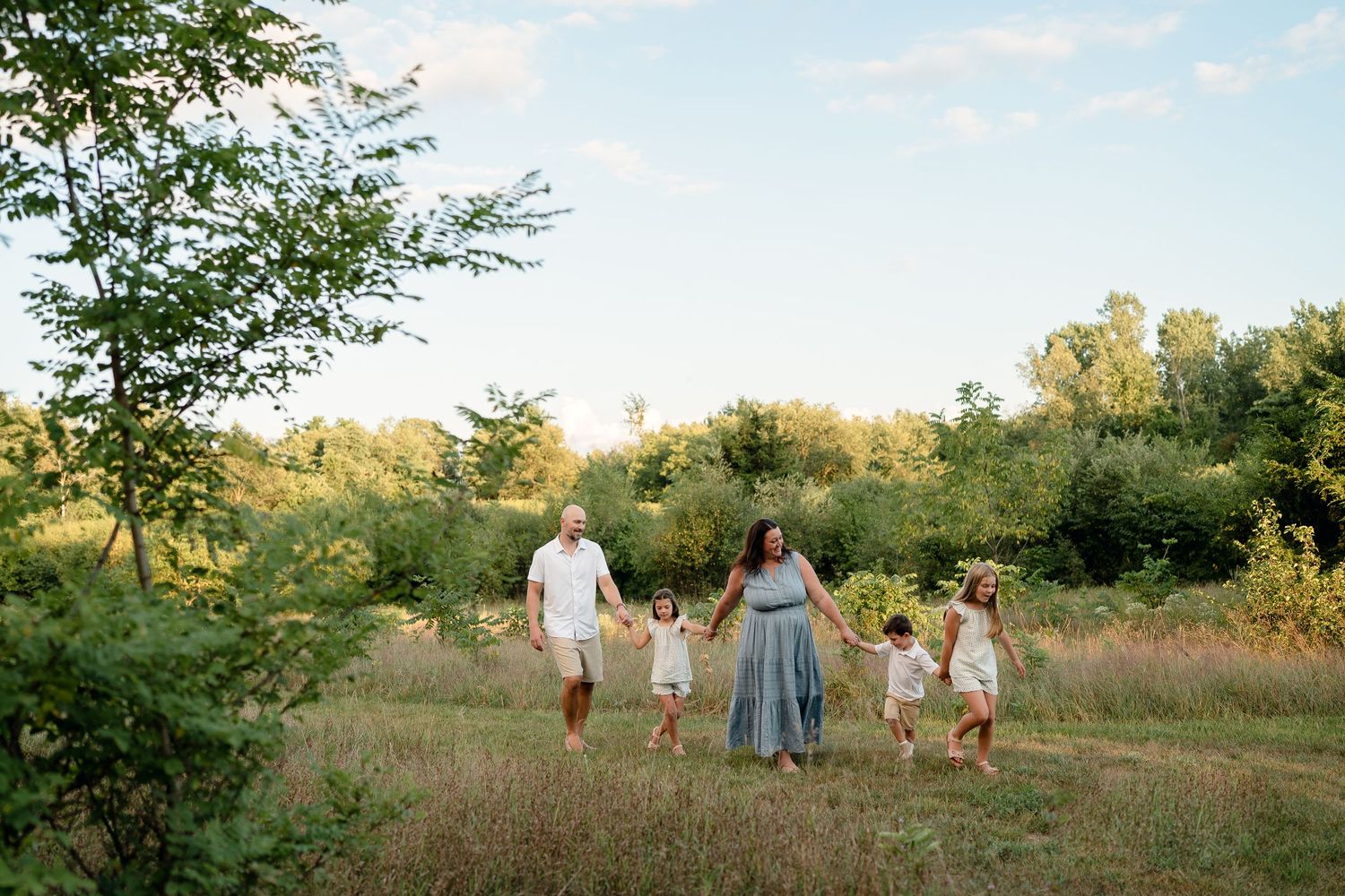Family of five holding hands walking through a sunny green meadow on a warm summer day.