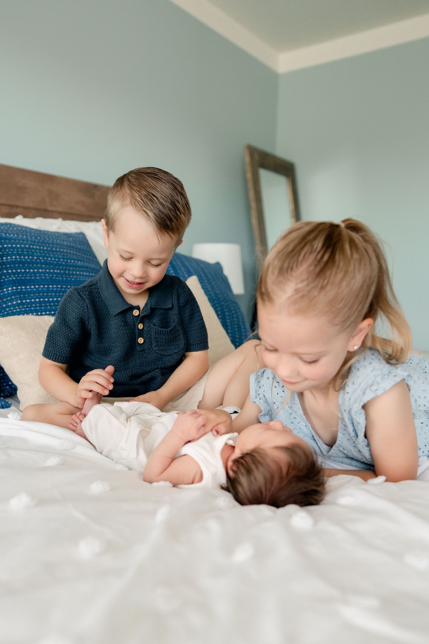 Two older siblings lovingly admire their newborn baby sibling on a white bed.