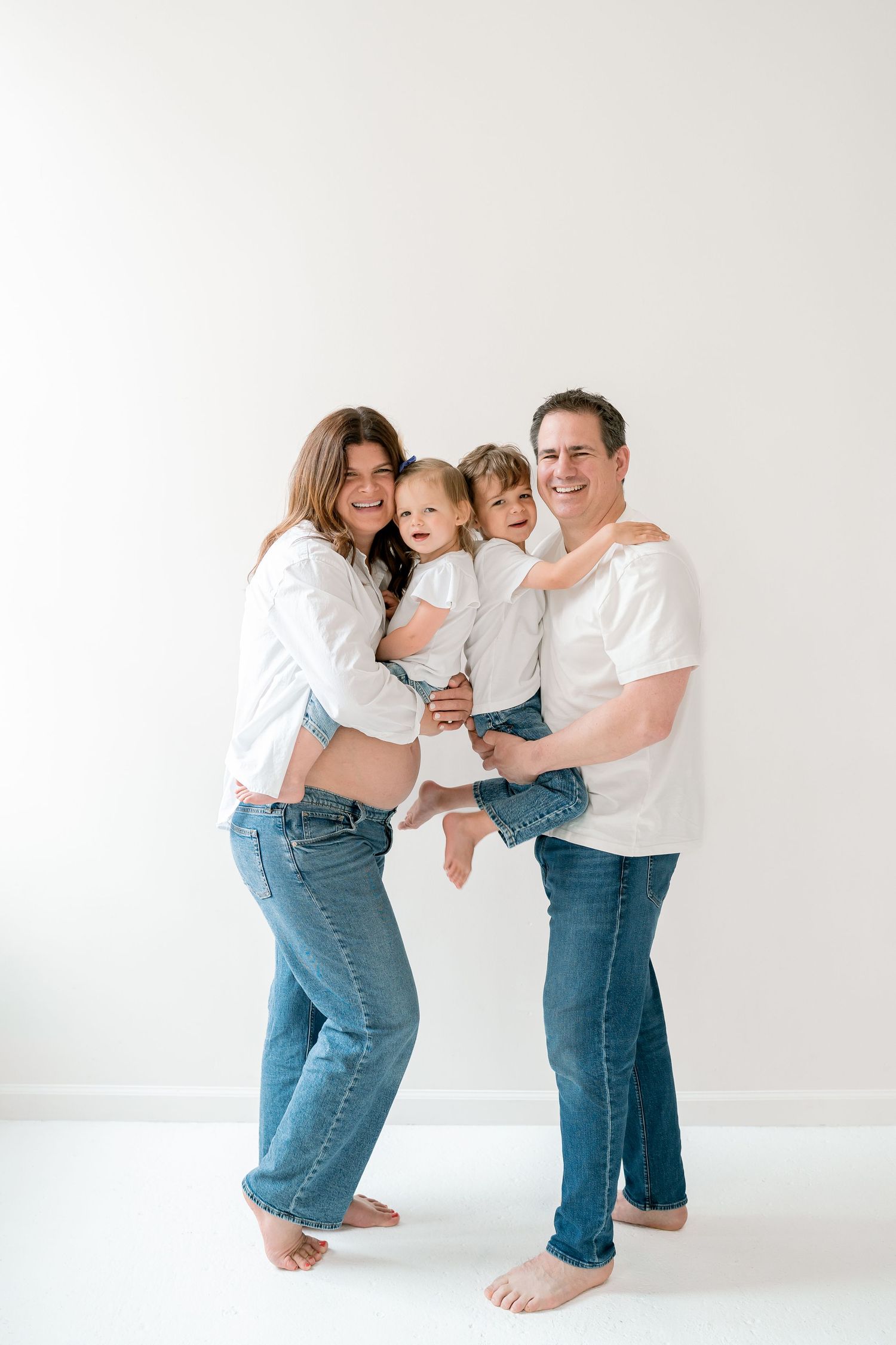 Joyful pregnant family in white tops and jeans, barefoot in a bright studio, parents holding their young son.