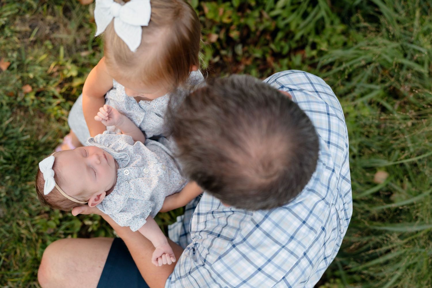 A father holds a newborn baby while a toddler girl with a white bow looks on, seated on grass outdoors.