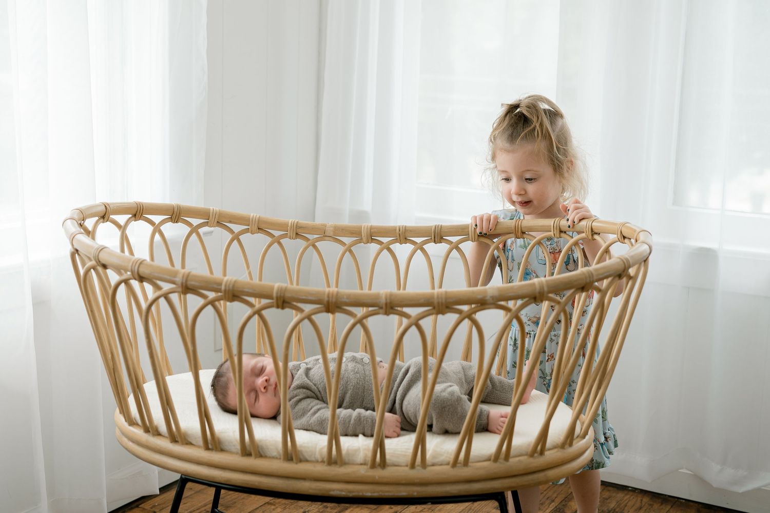 Toddler girl smiling at newborn baby lying in a natural rattan bassinet near white curtains.