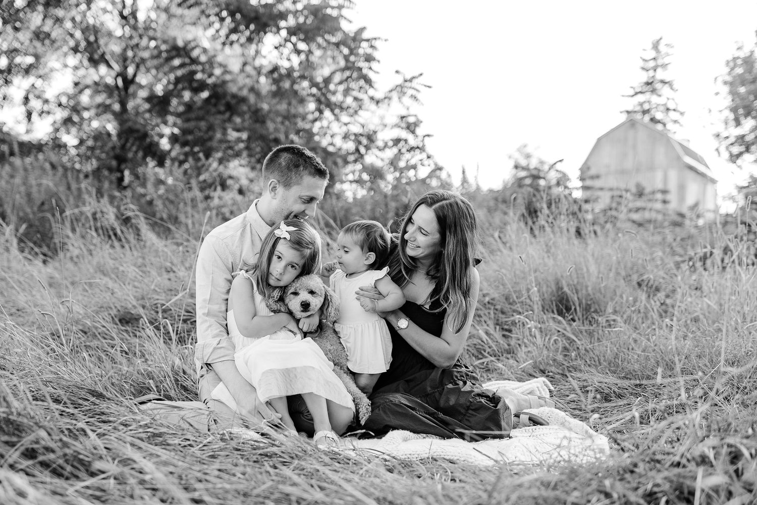 Black and white photo of family sitting together in a grassy field near a barn.