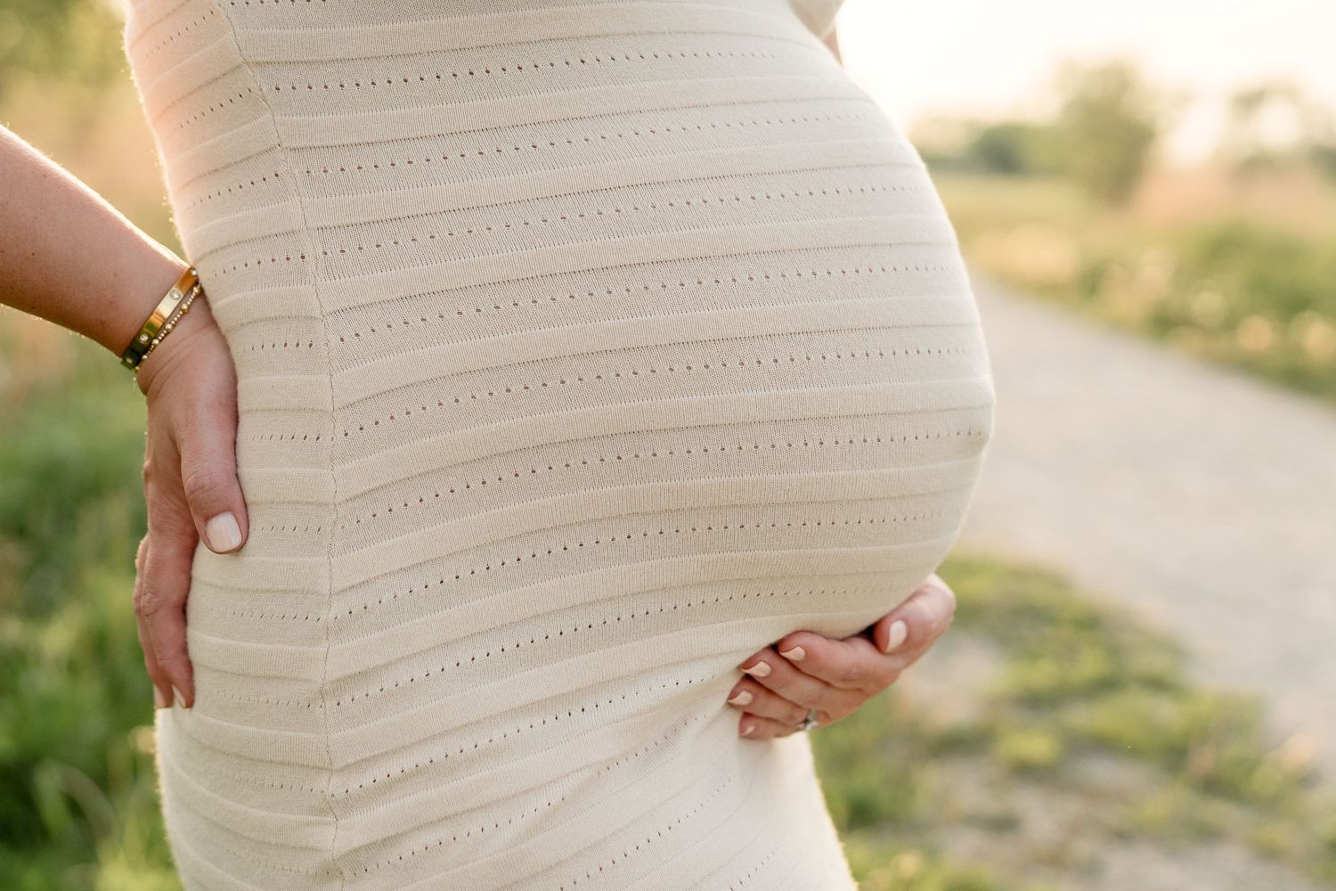 Close-up of pregnant belly in cream ribbed dress with hands gently cradling bump outdoors.